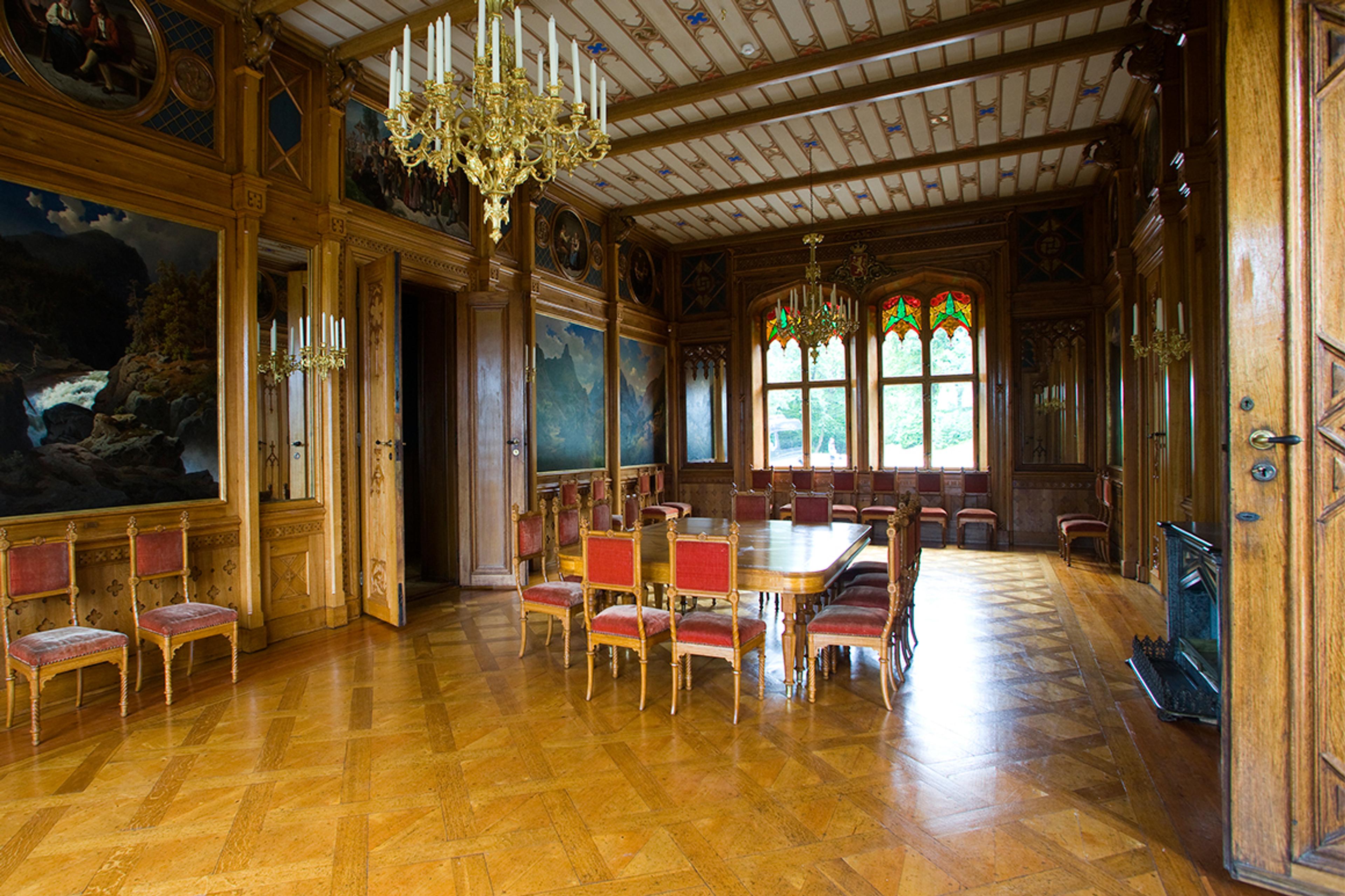 Bord og stoler står midt i et rom med store vinduer og vegger utsmykket med malerier.