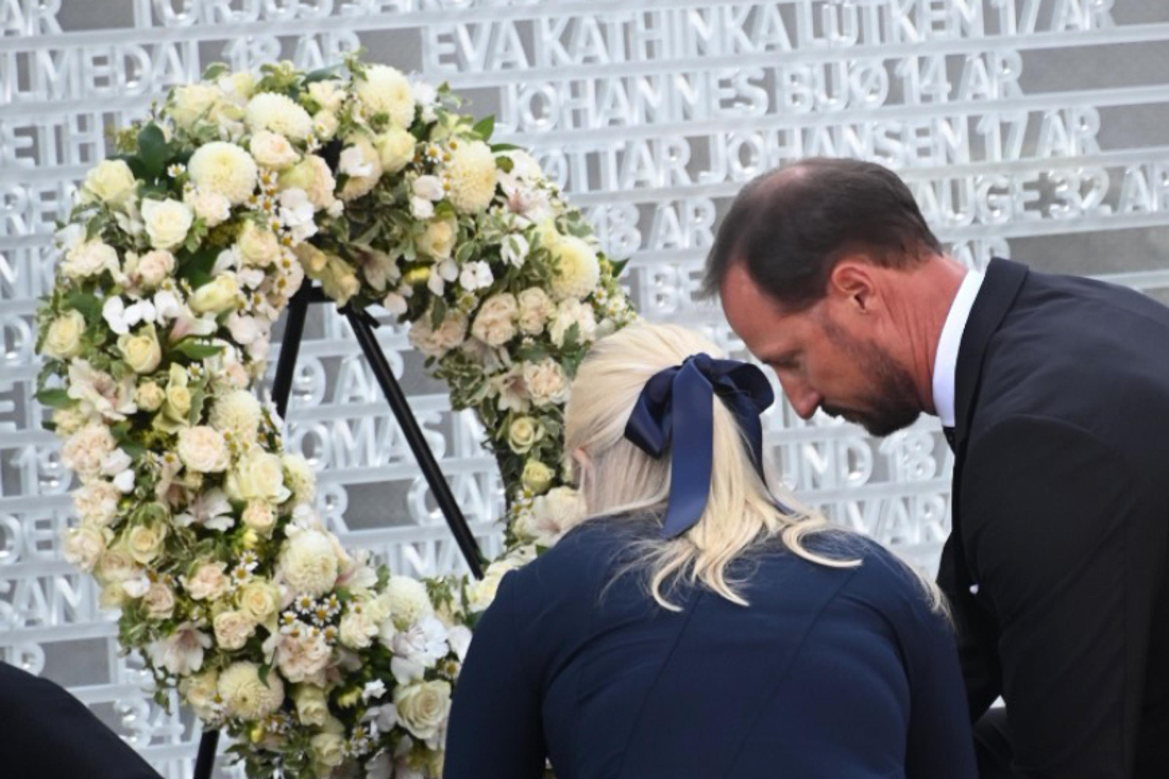 Kronprinsparet sitter på huk foran en stor krans med hvite blomster og tavlen med navnene.