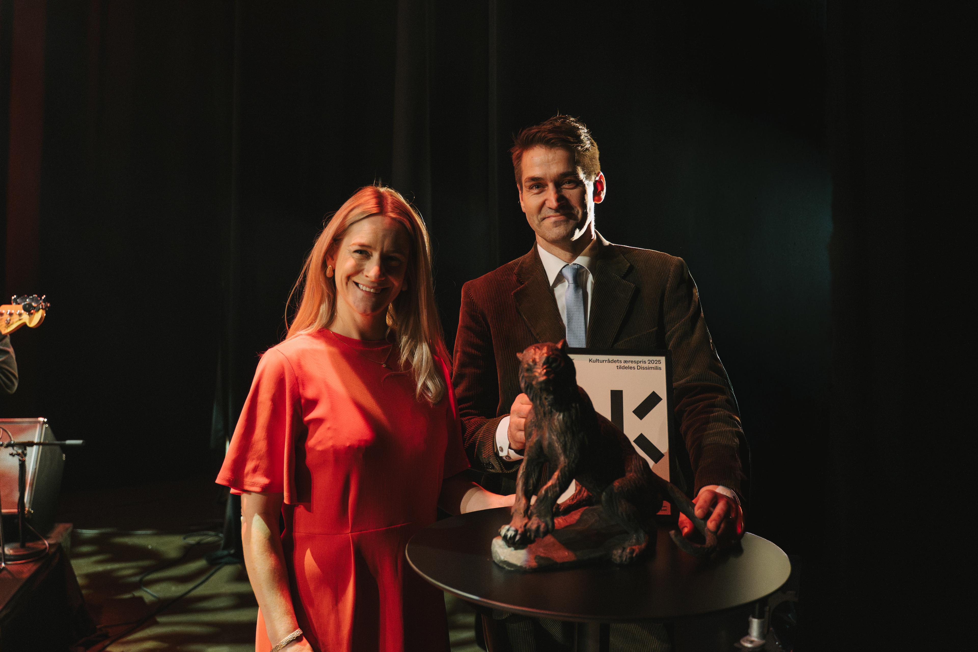 Nathalie Sturim og Sigmund Løvåsen står side om side og ser inn i kamera. På eit rundt ståbord framfor dei står ein løvestatuett.
