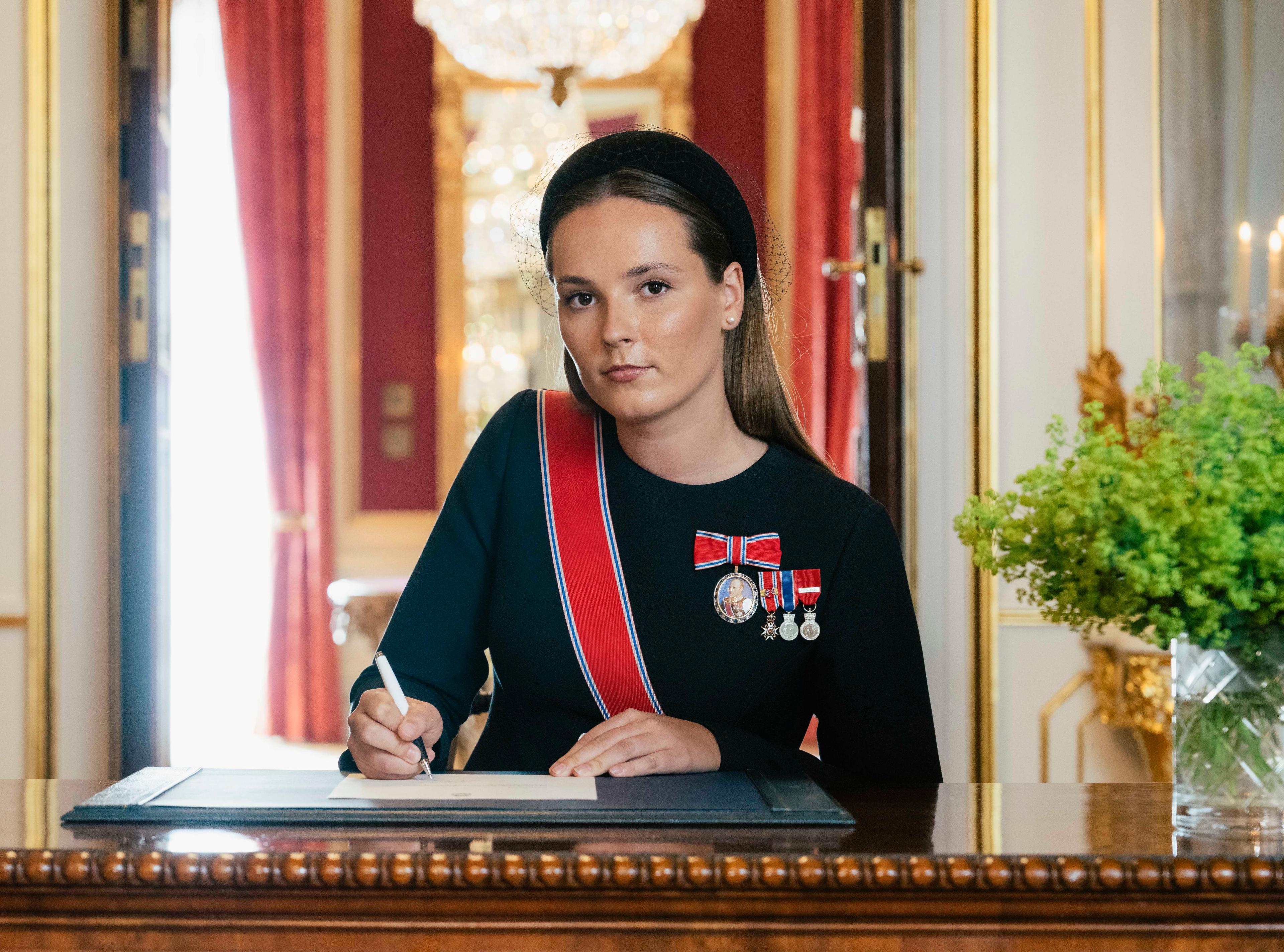 Kronprinsessen sitter ved et skrivebord i sort kjole med ordener og medaljer. Hun holder en penn i hånden og ser opp, inn i kameraet.
