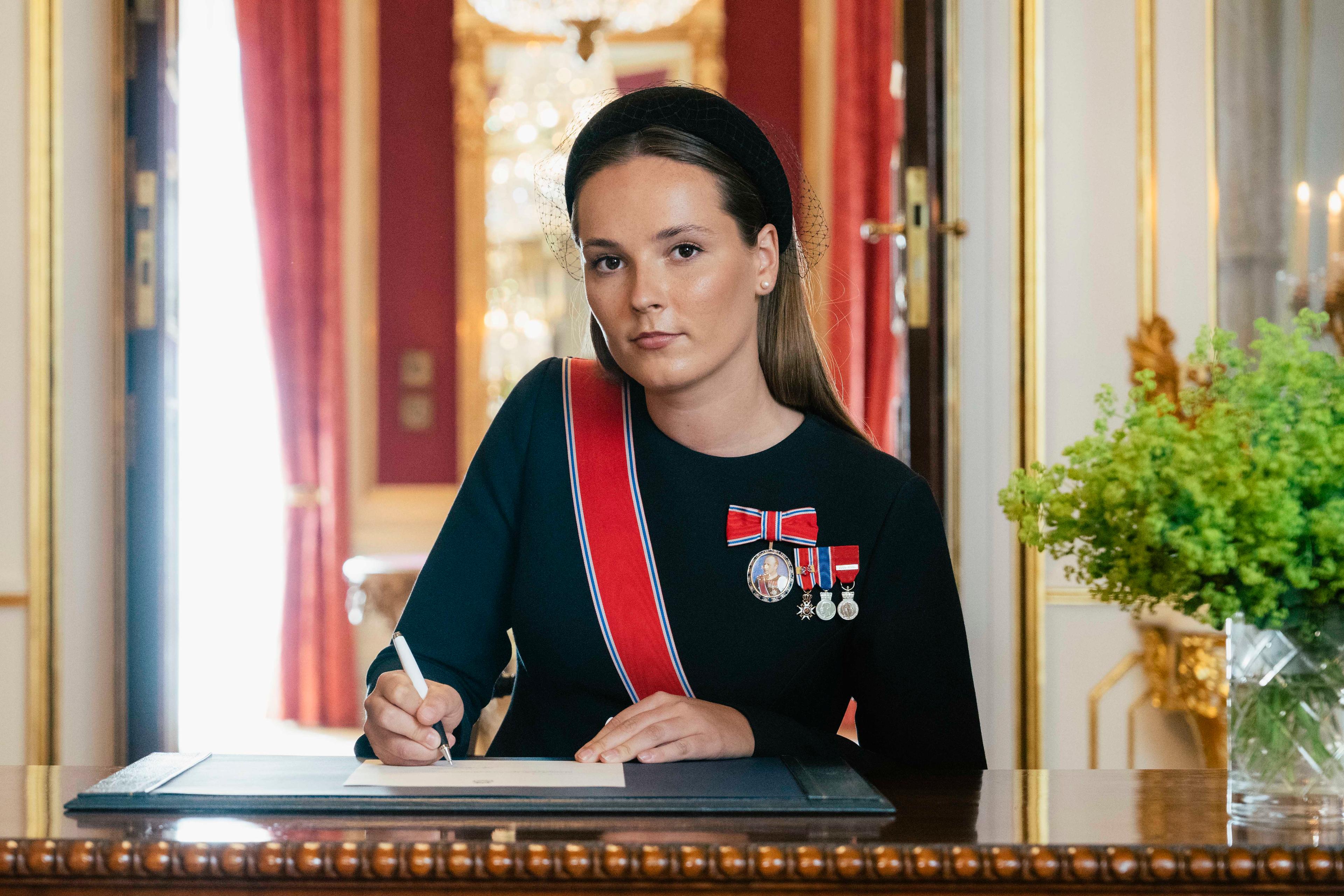 Kronprinsessen sitter ved et skrivebord i sort kjole med ordener og medaljer. Hun holder en penn i hånden og ser opp, inn i kameraet.