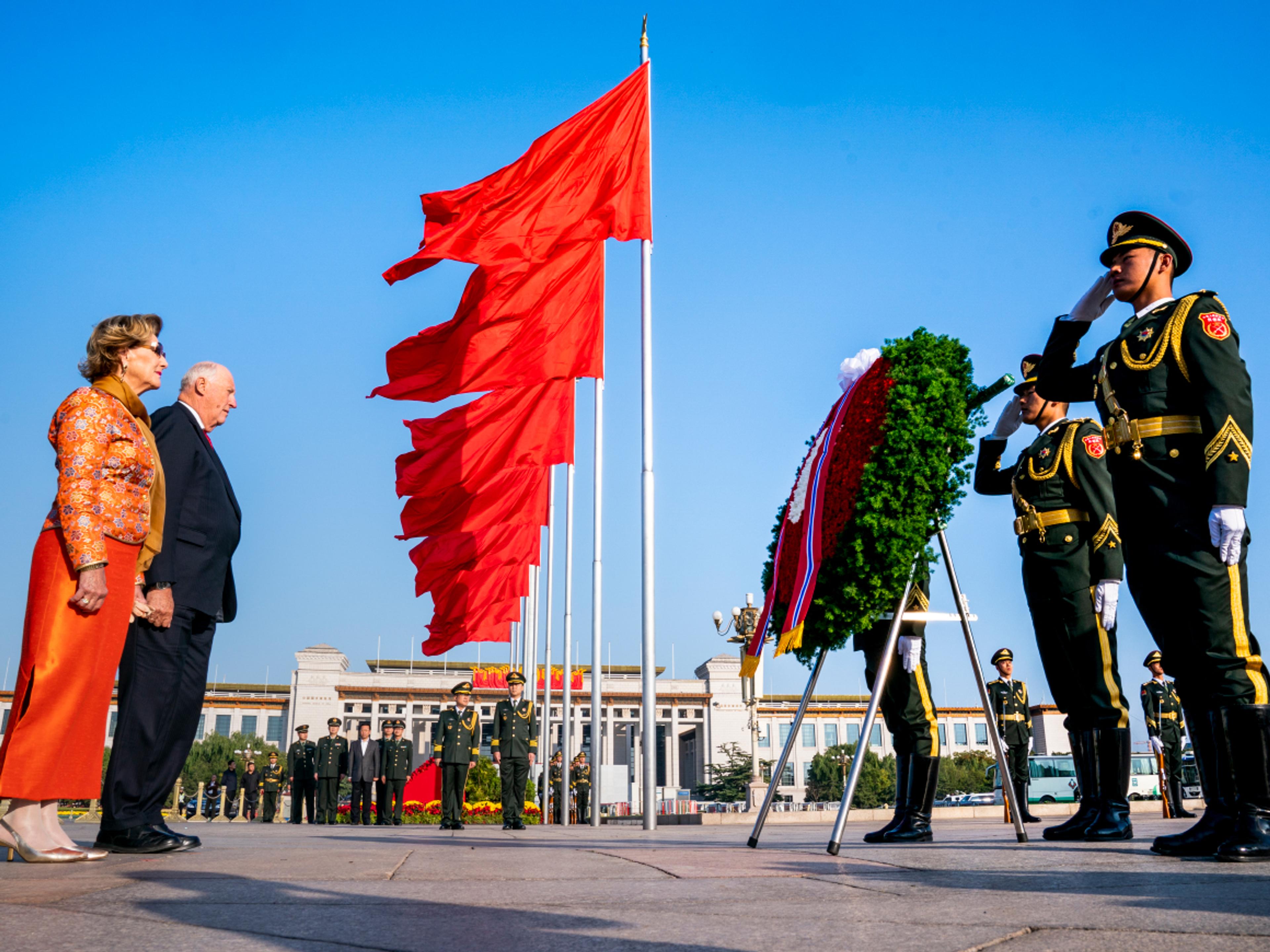 Kongeparet står høytidelig foran en stor blomsterkrans, med æresvakt på hver side. En lang rekke røde, kinesiske flagg vaier.