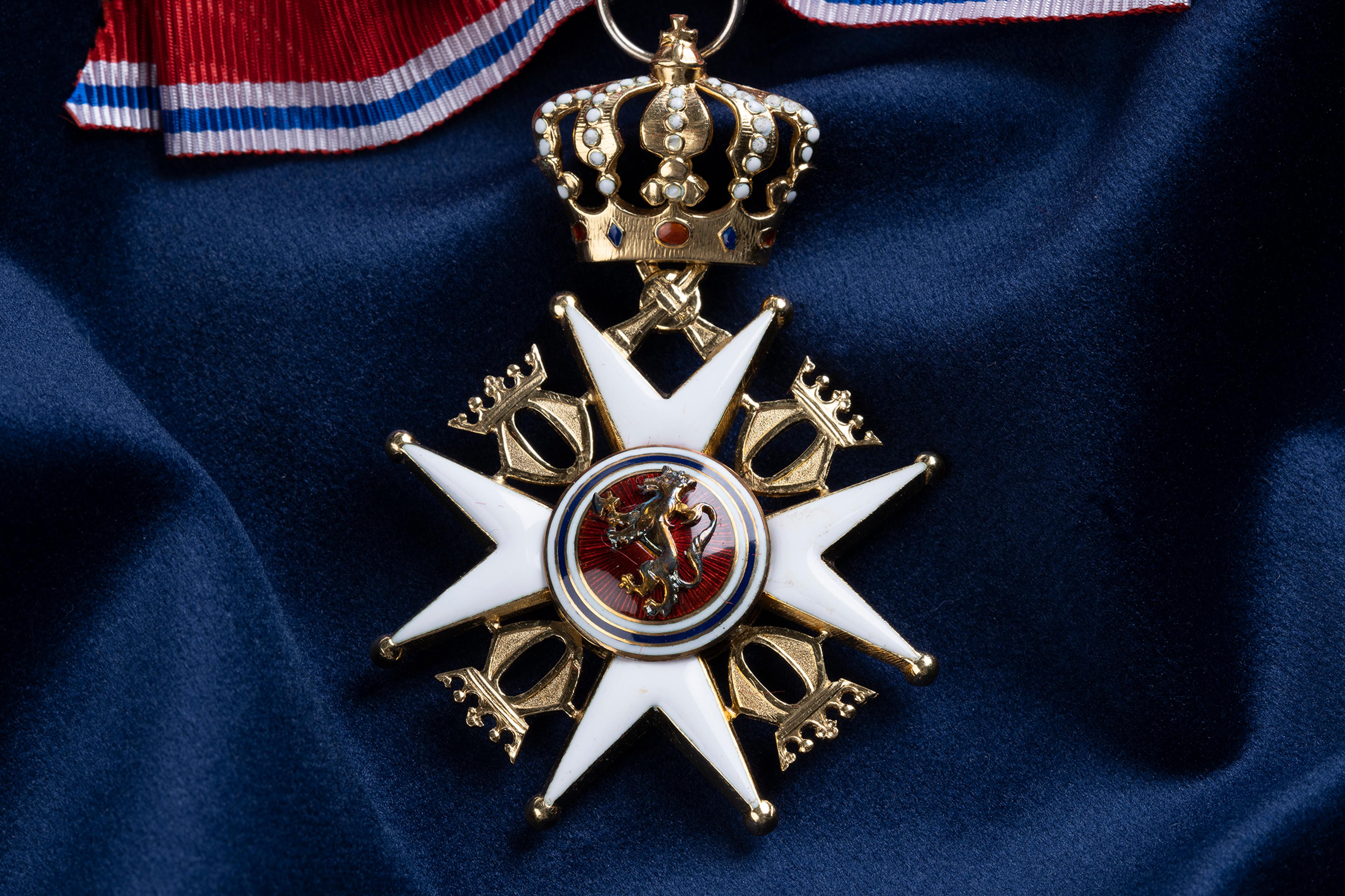 Nærbilde av ordensmerket festet i rødt bånd med blå og hvite striper på en bakgrunn av blått klede.