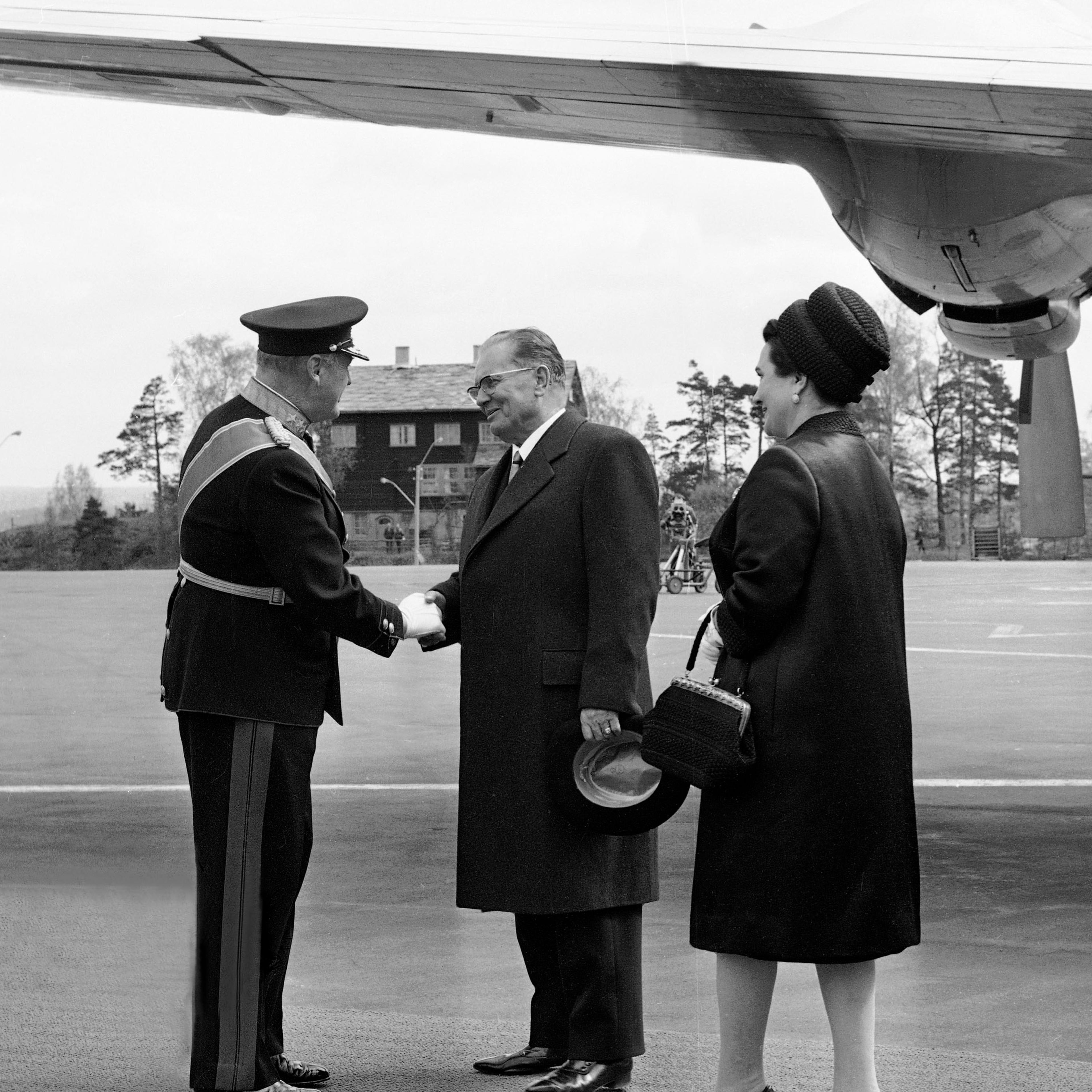 Sort-hvitt foto av Kong Olav i uniform, som håndhilser på "Tito" på rullebanen på Fornebu.