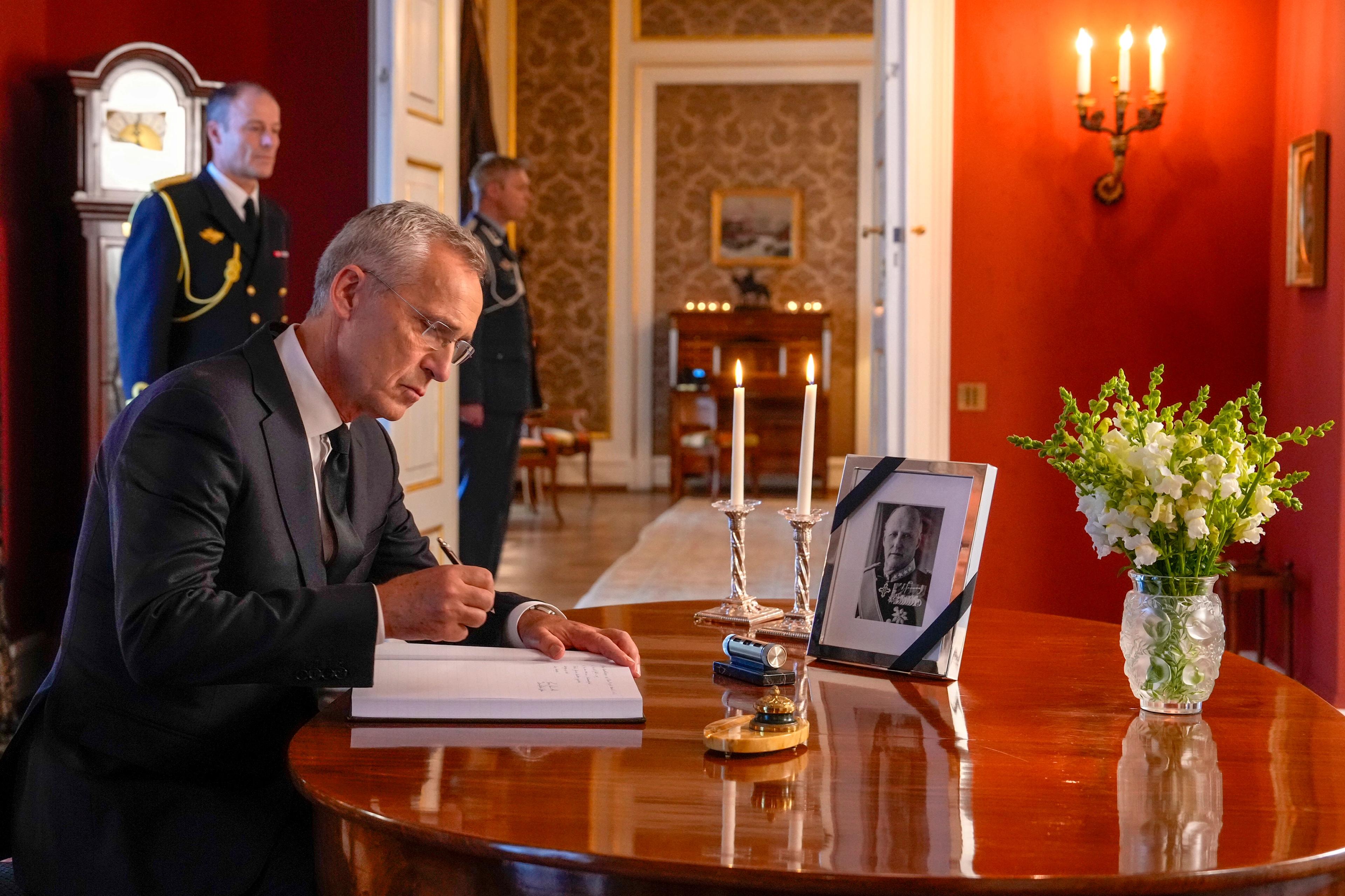 Stoltenberg sitter ved et bord og skriver. På bordet står et bilde av Kong Harald med sørgeflor omkring rammen og en bukett hvite blomster.