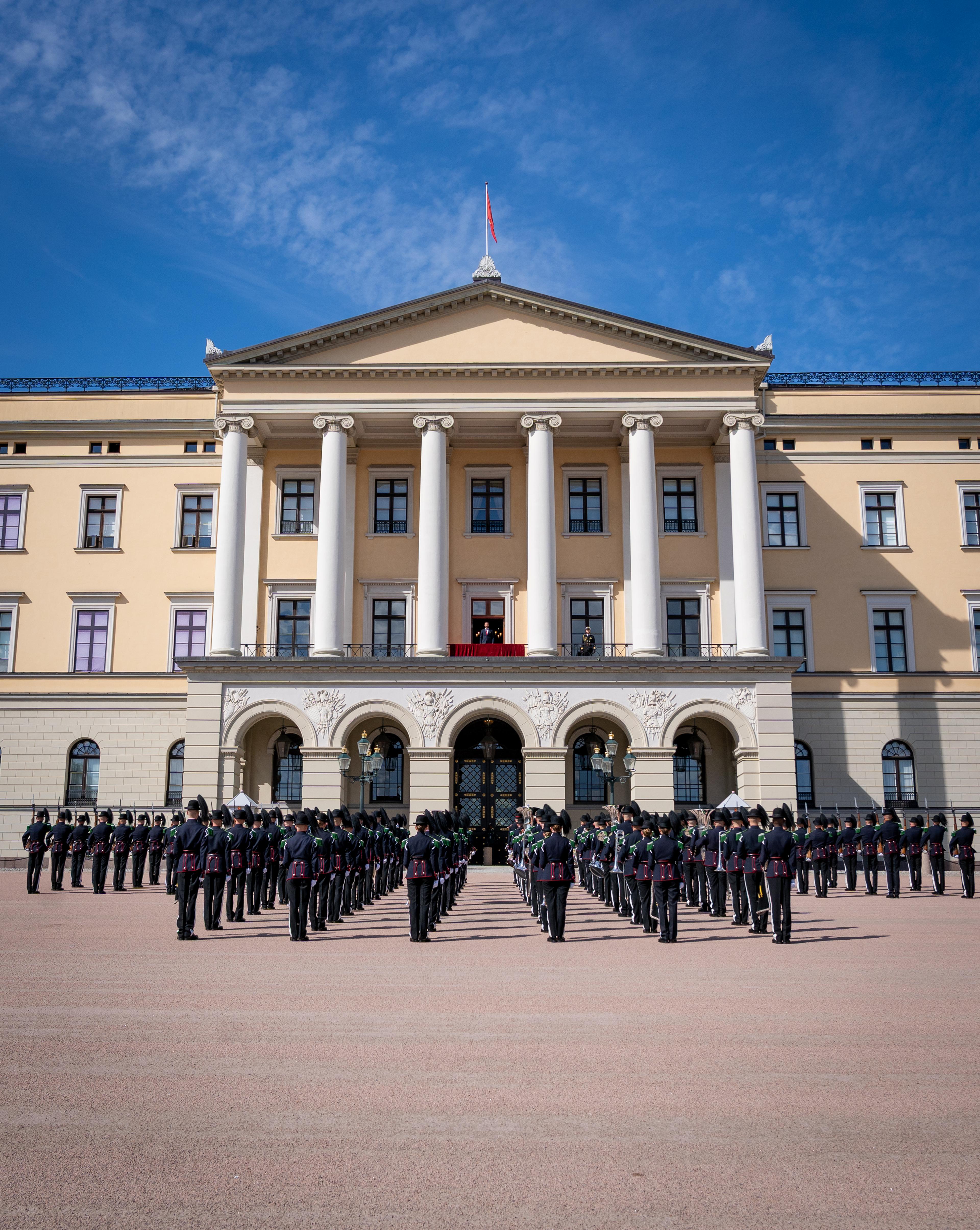 Musikk- og Drilltroppen står oppstilt foran Slottet. På Slottsbalkongen står Kronprins Haakon.