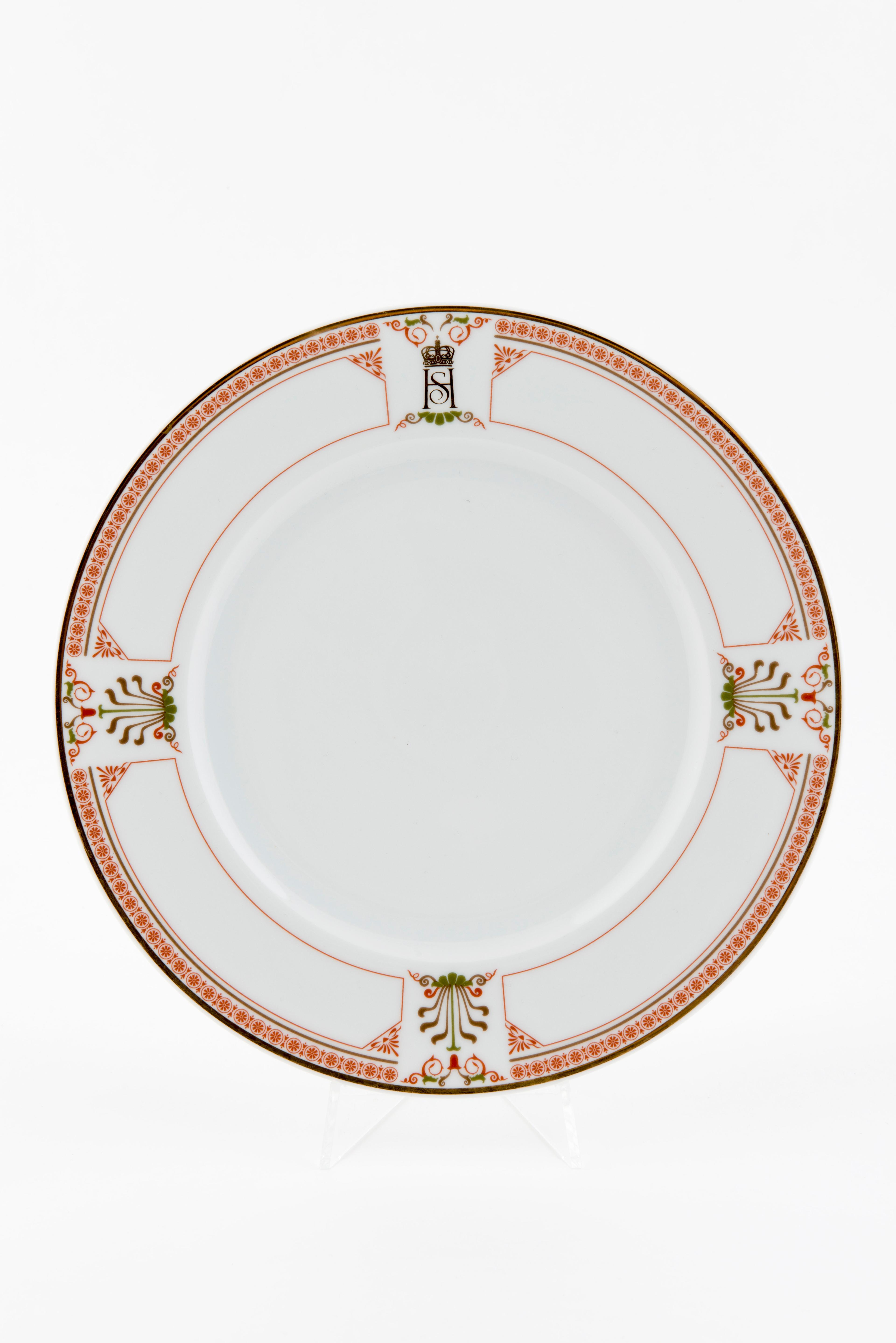 En hvit tallerken med en smal kant med rød-orange dekor. Tallerkenkanten har også tre felt med større palmetter i tillegg til Kongeparets fellesmonogram.
