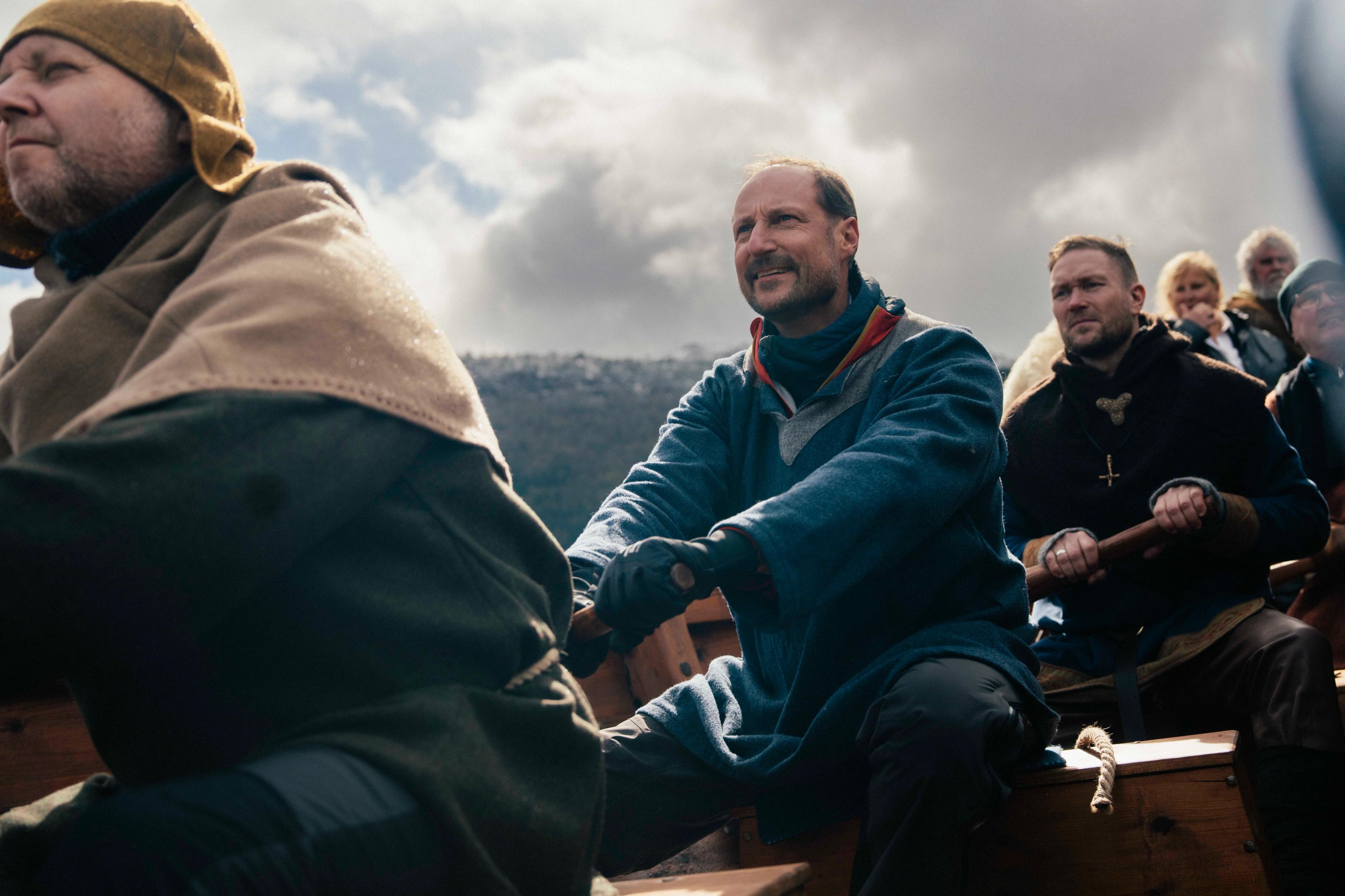 Kronprins Haakon ror sammen med resten av mannskapet i Myklebustskipet. I bakgrunnen ser man fjell og mørke skyer