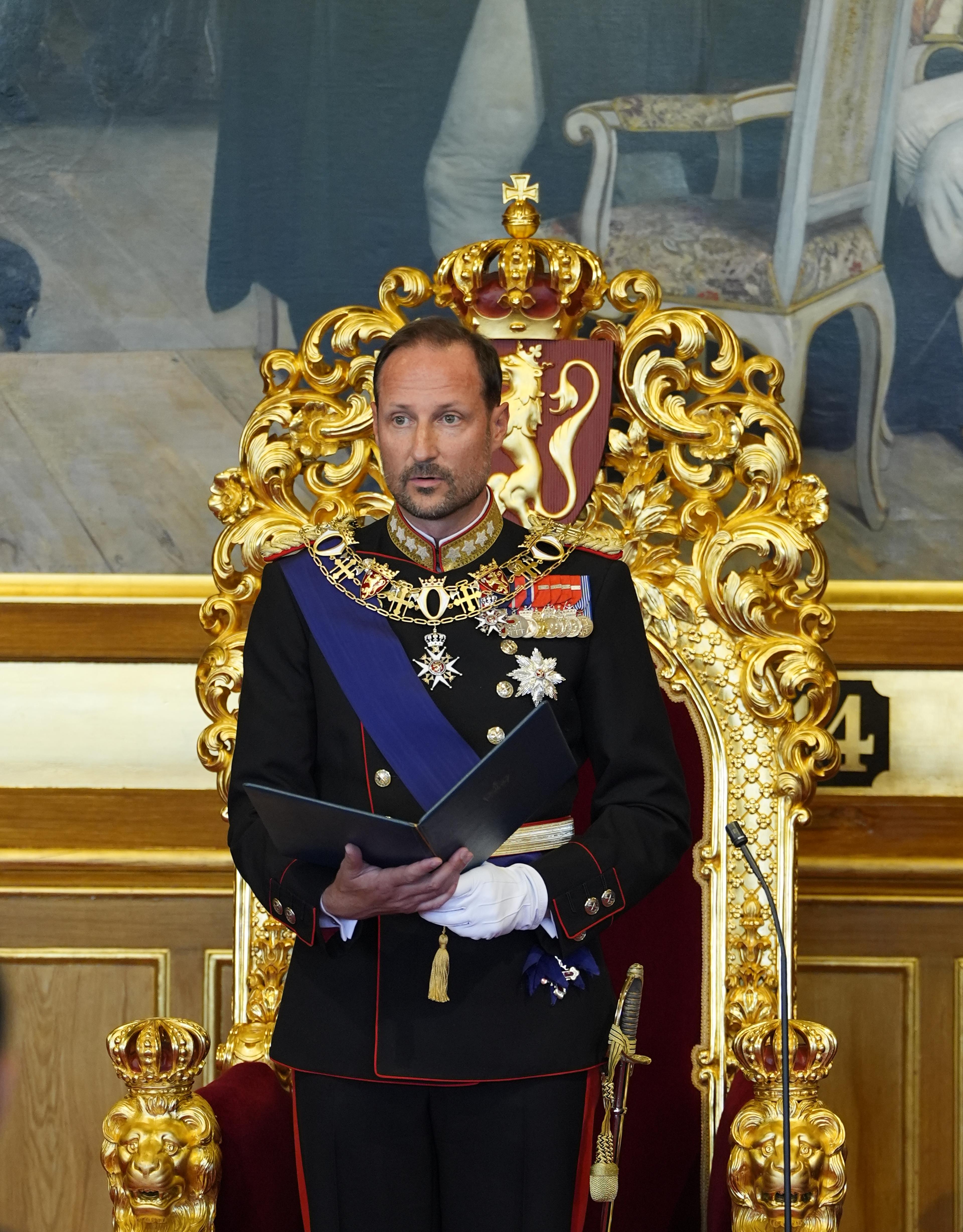 Kong Haakon står foran tronen i Stortingssalen. Han er i gallauniform og holder en mappe med teksten i hendene.