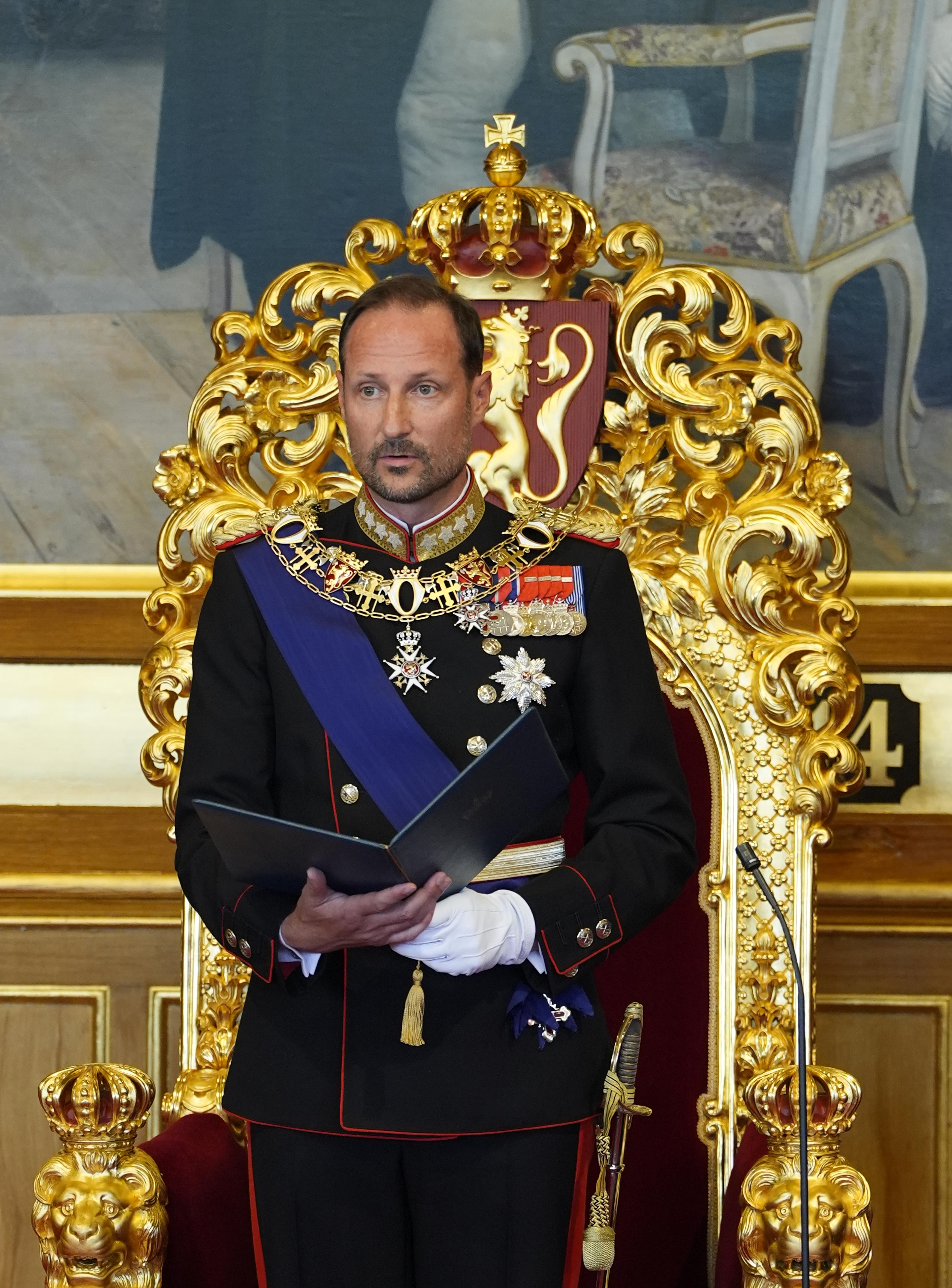 Kong Haakon står foran tronen i Stortingssalen. Han er i gallauniform og holder en mappe med teksten i hendene.