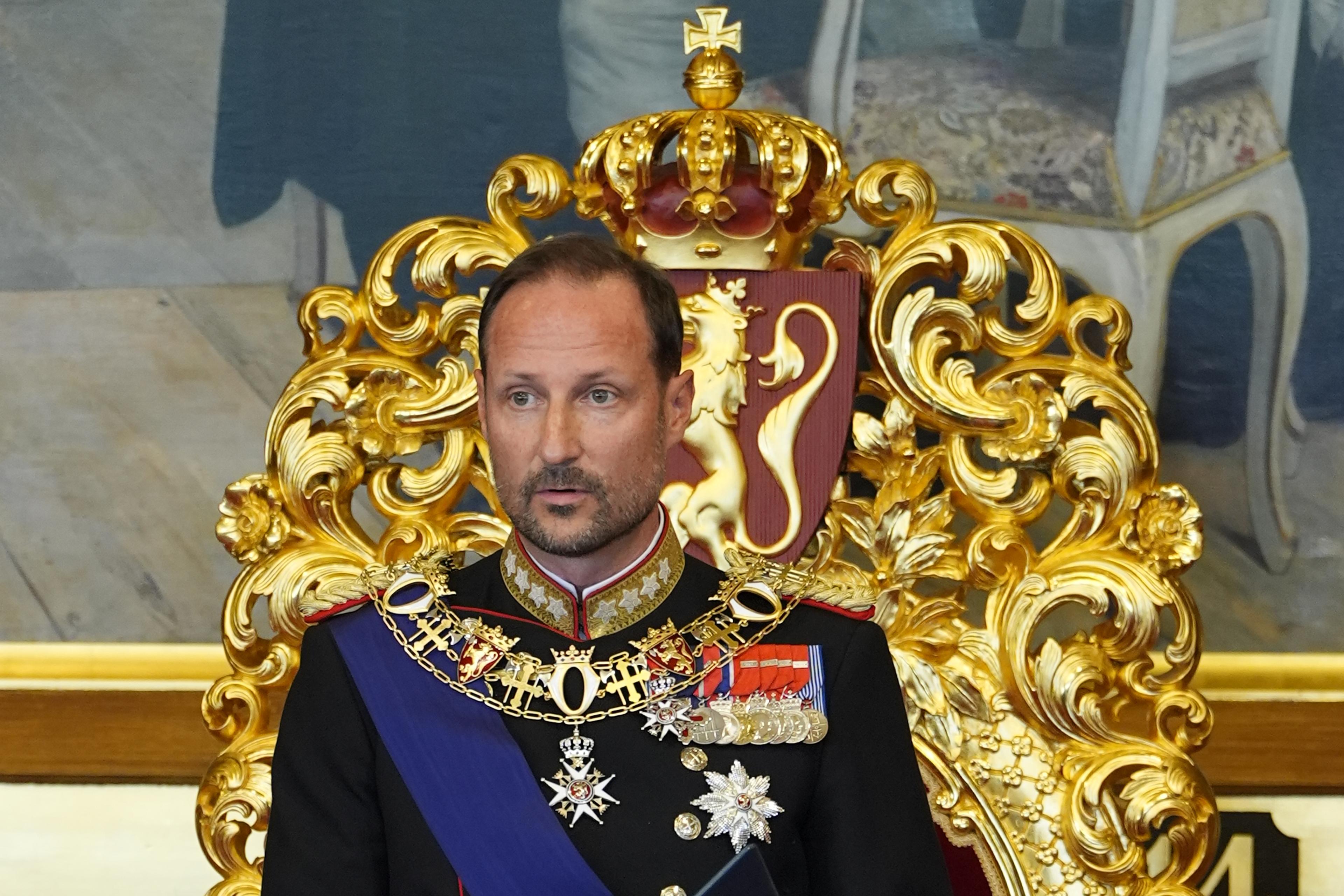 Kong Haakon står foran tronen i Stortingssalen. Han er i gallauniform og holder en mappe med teksten i hendene.