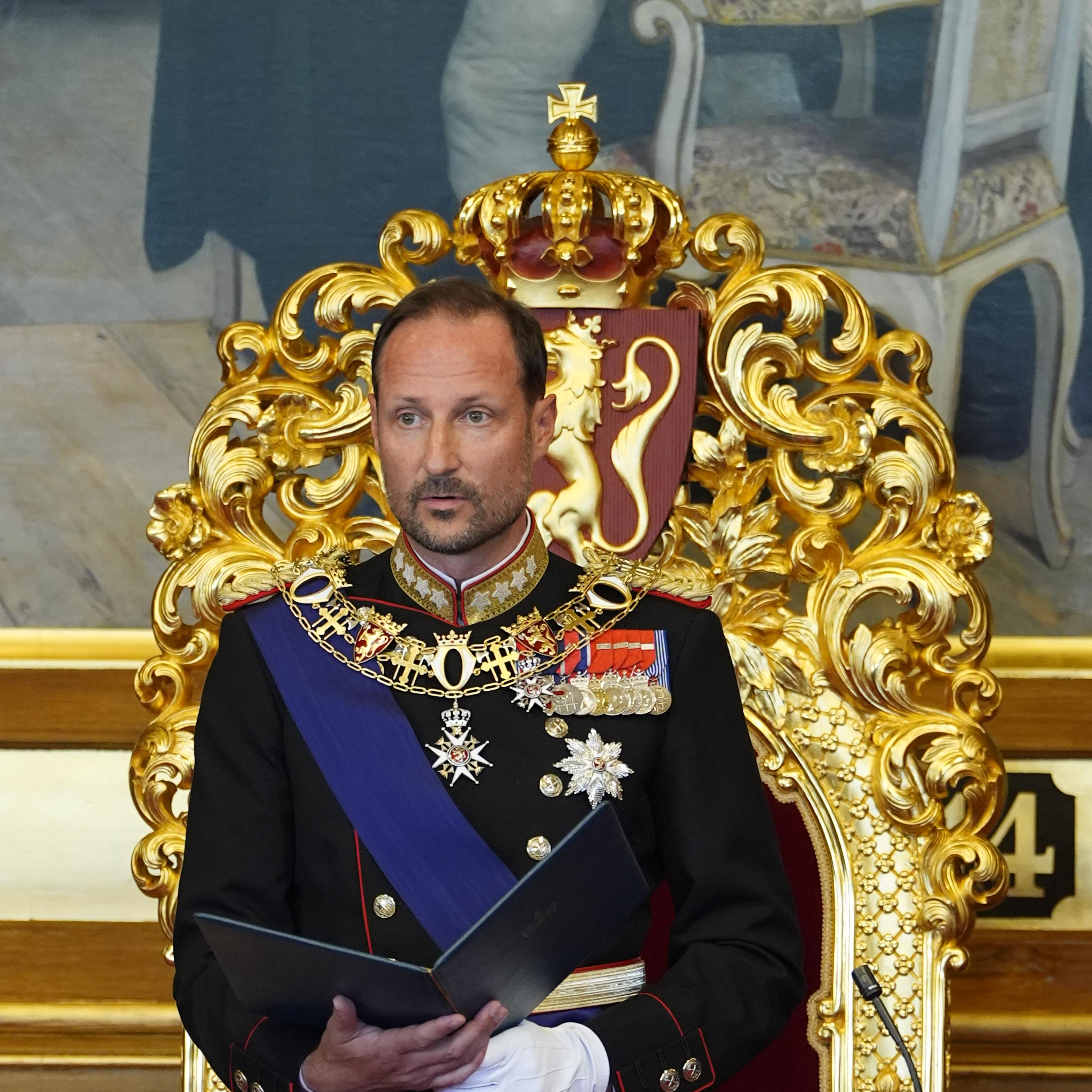 Kong Haakon står foran tronen i Stortingssalen. Han er i gallauniform og holder en mappe med teksten i hendene.
