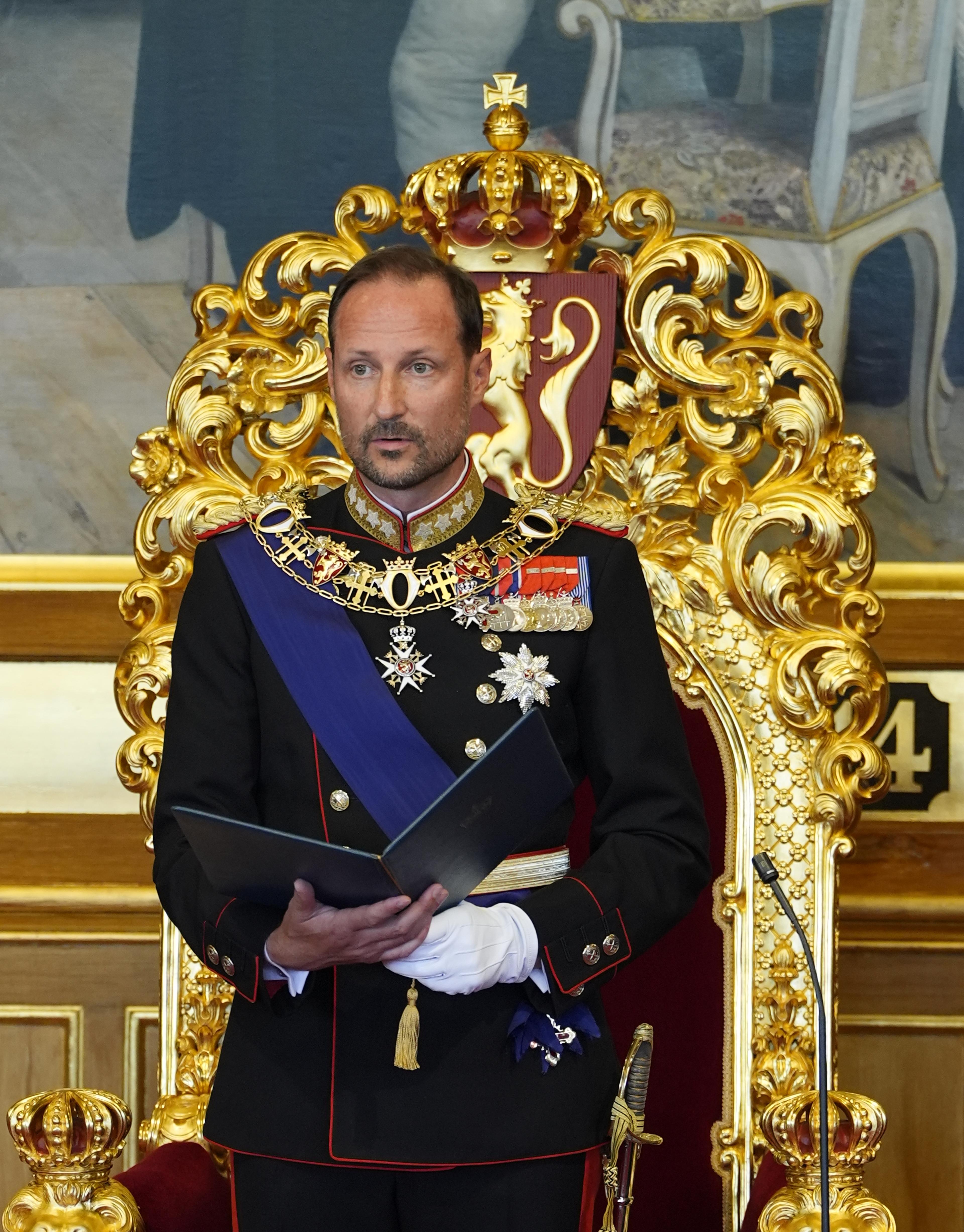 Kong Haakon står foran tronen i Stortingssalen. Han er i gallauniform og holder en mappe med teksten i hendene.