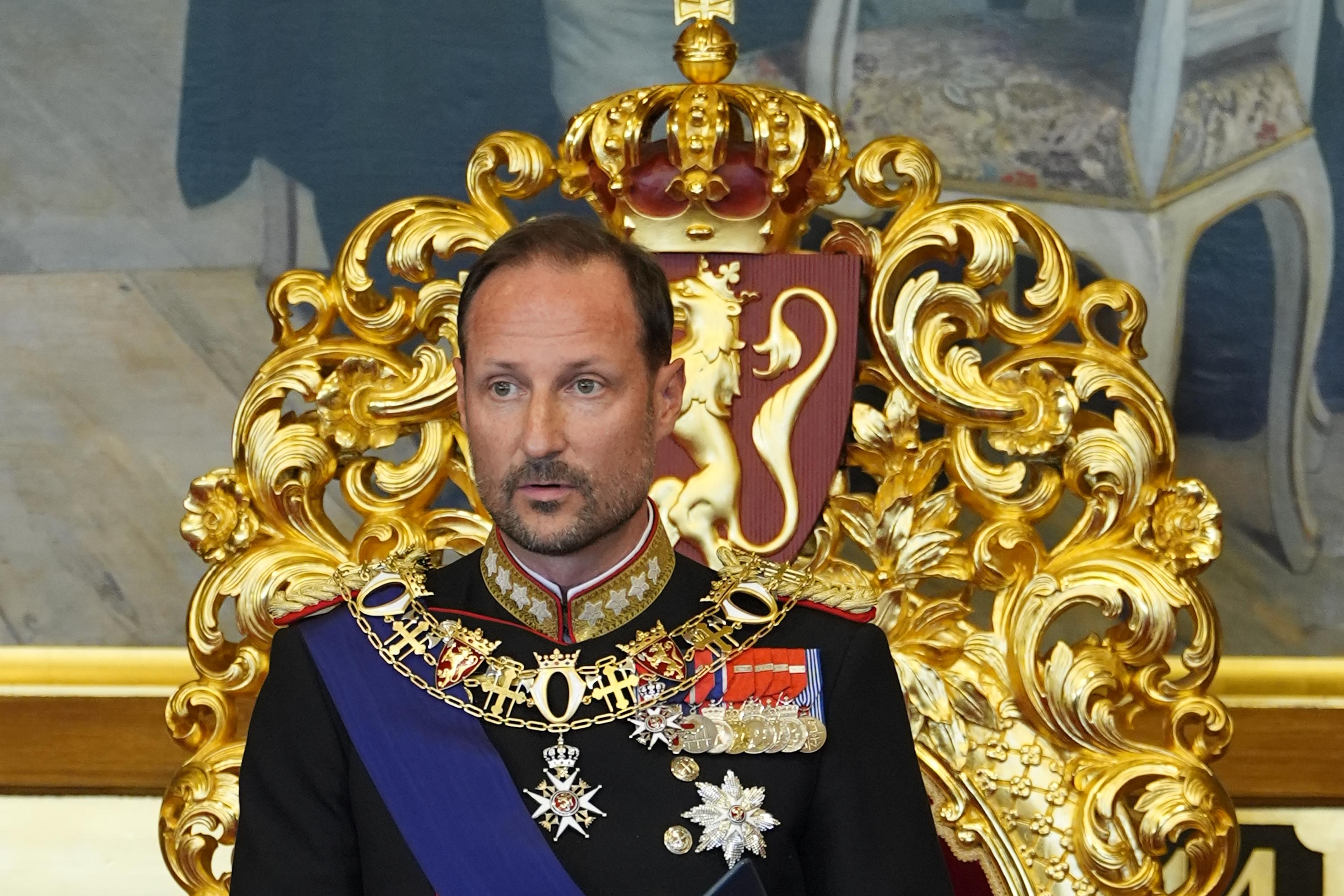 Kong Haakon står foran tronen i Stortingssalen. Han er i gallauniform og holder en mappe med teksten i hendene.