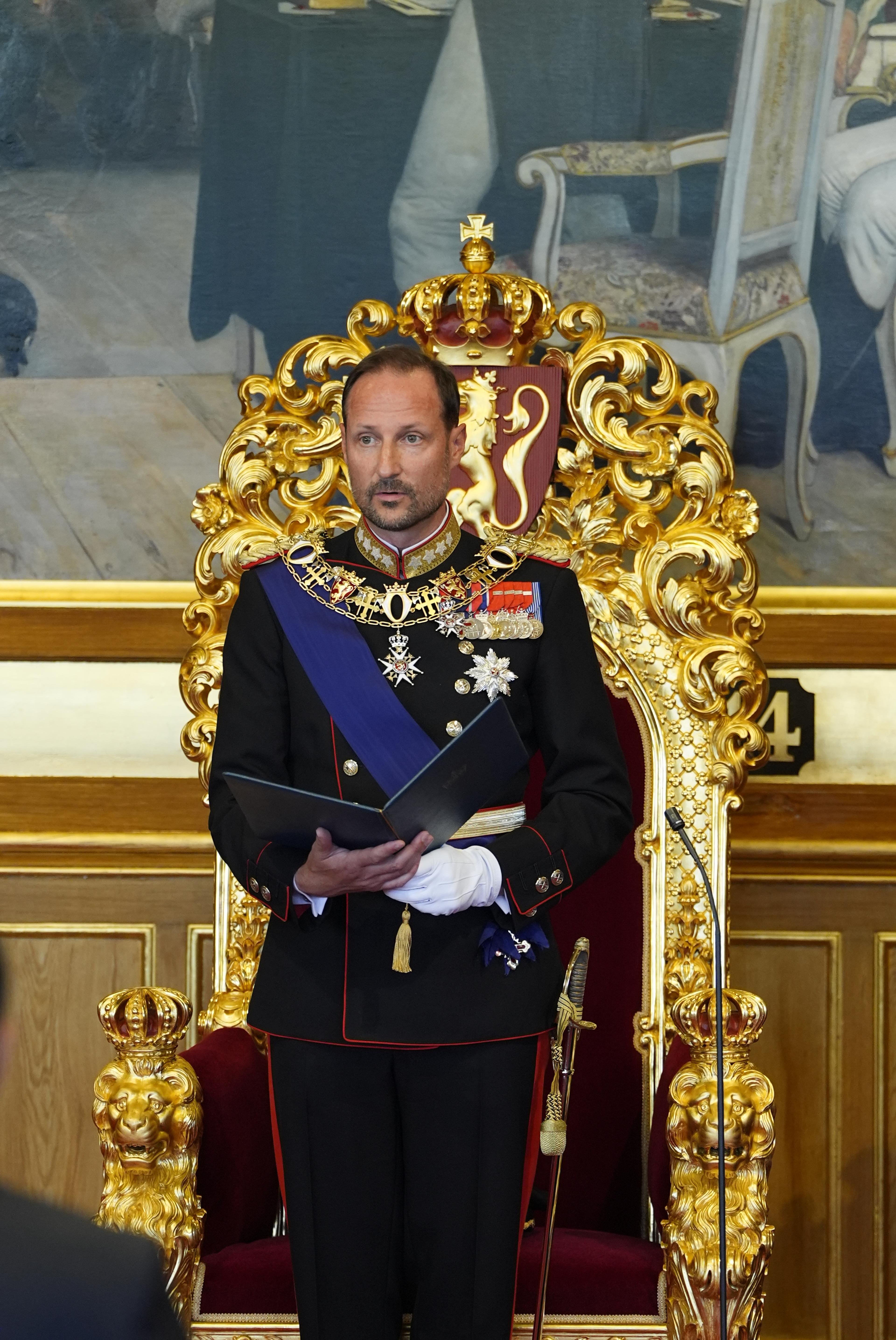 Kong Haakon står foran tronen i Stortingssalen. Han er i gallauniform og holder en mappe med teksten i hendene.