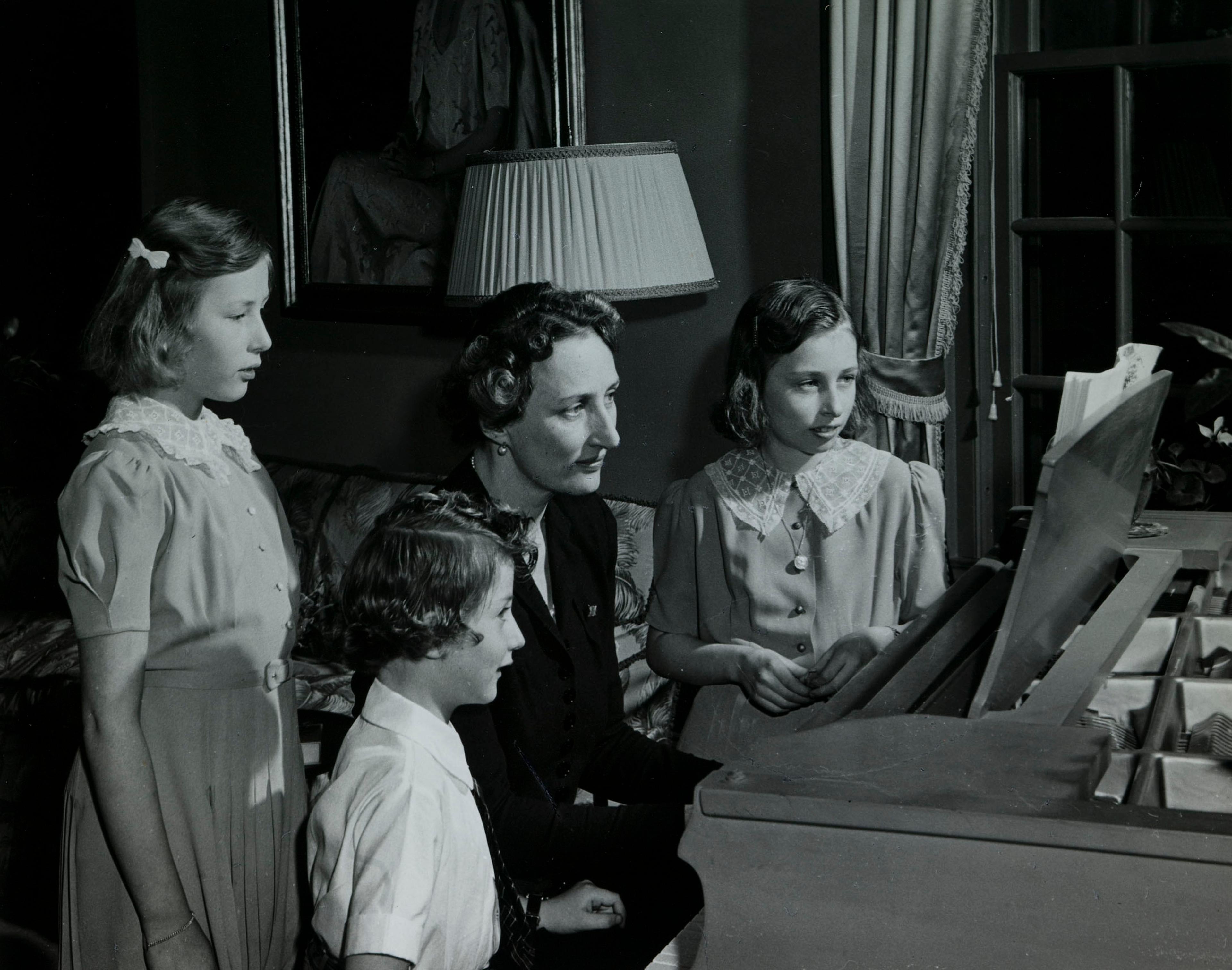 Kronprinsesse Märtha sitter på pianokrakken. Barna står rundt og ser på notearket.