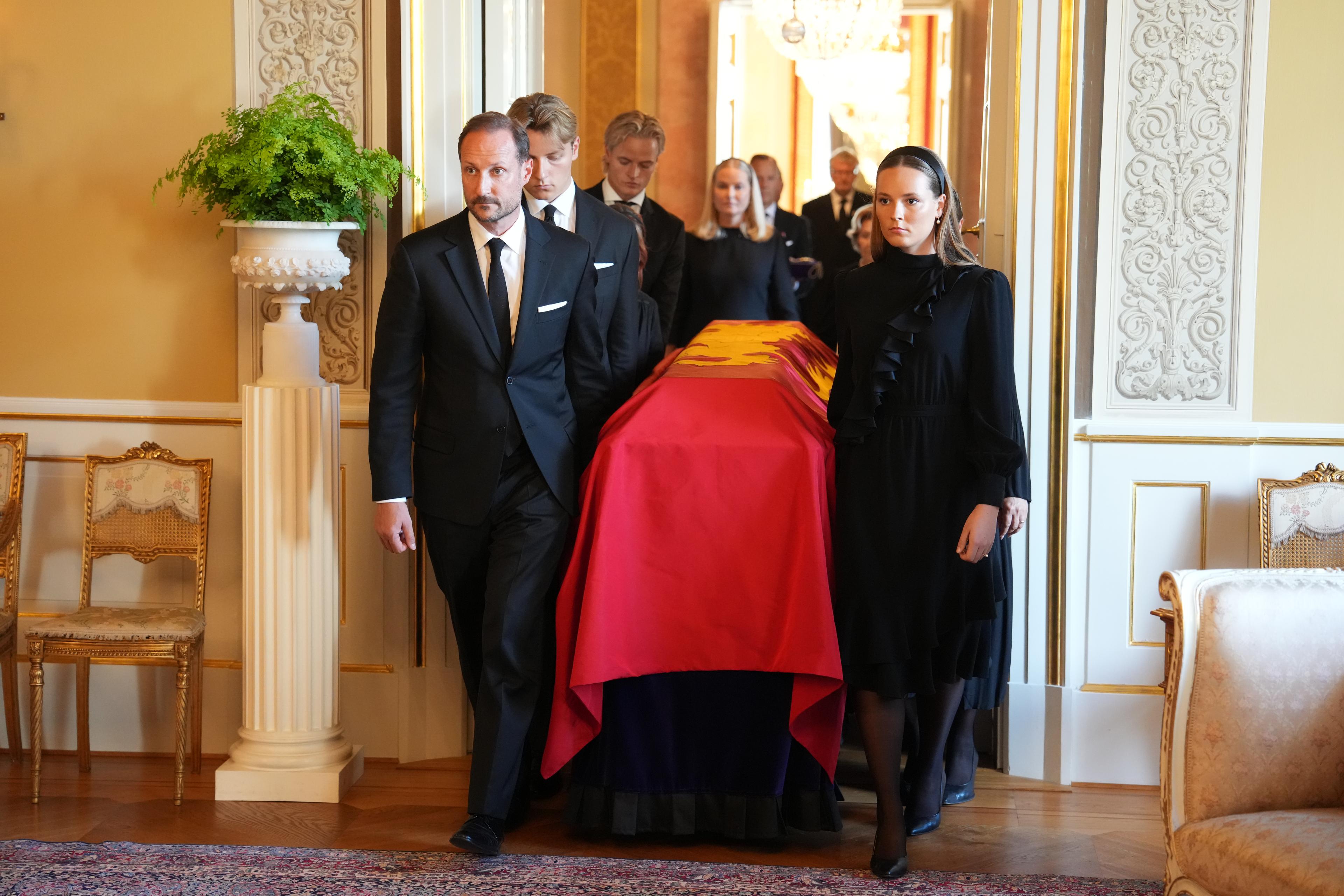 Båra er drapert i kongeflagget og står på ein katafalk. I spissen for prosesjonen går Kong Haakon og Kronprinsesse Ingrid Alexandra.
