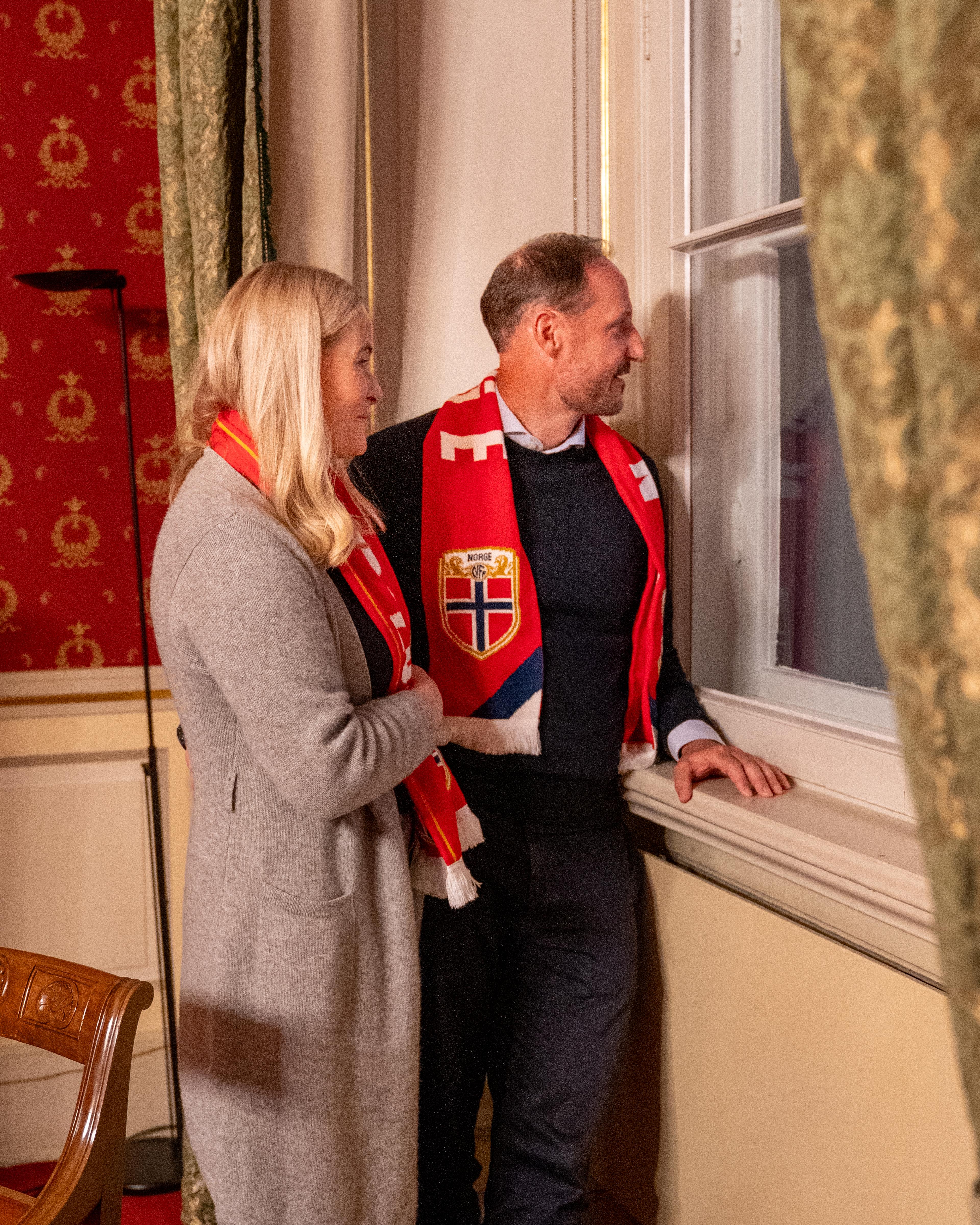 Kronprinsparet er iført supporterskjerf og ser ut av vinduet på Slottet.