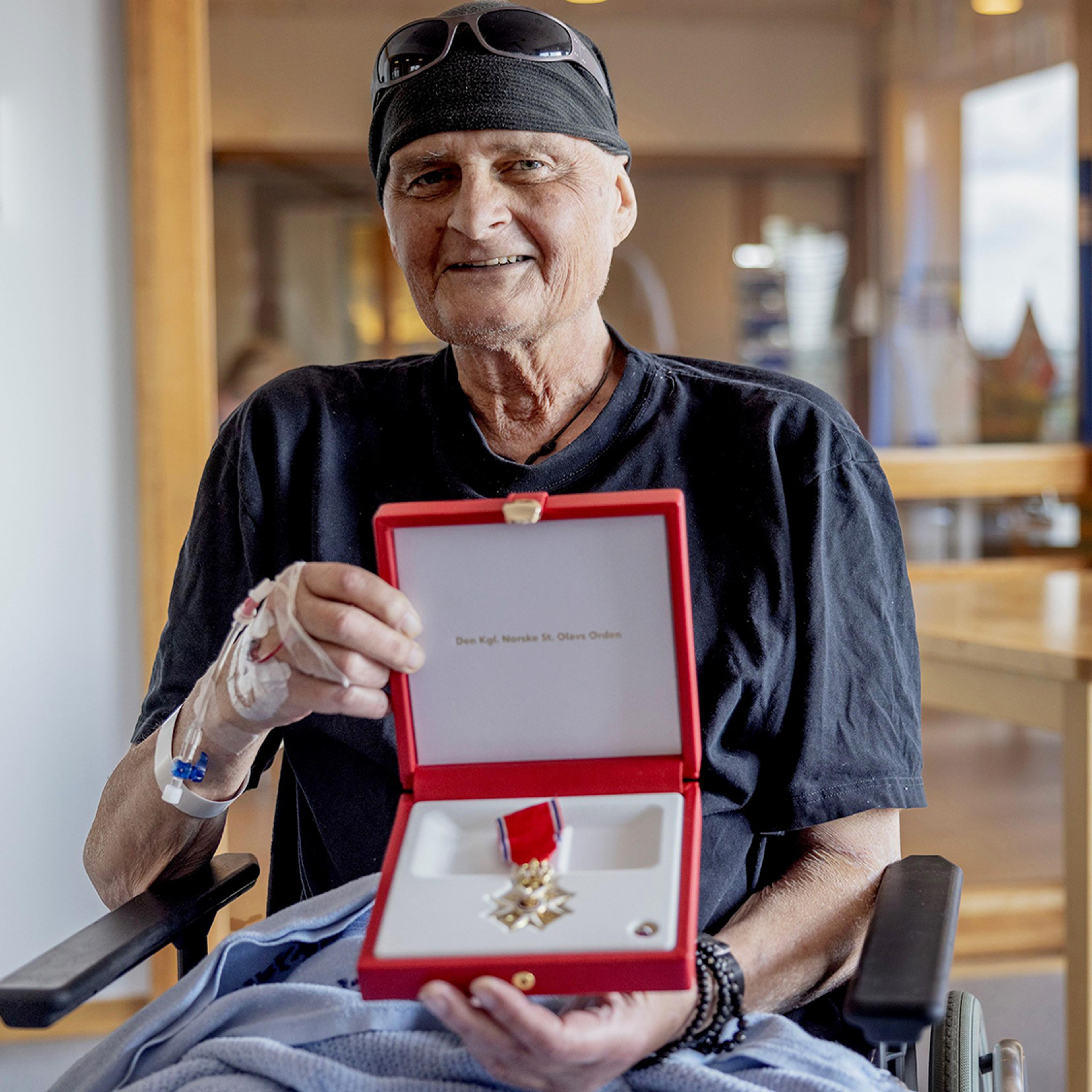 En smilende Henden sitter i på sykehuset og viser fram skrinet med St. Olavs Orden.