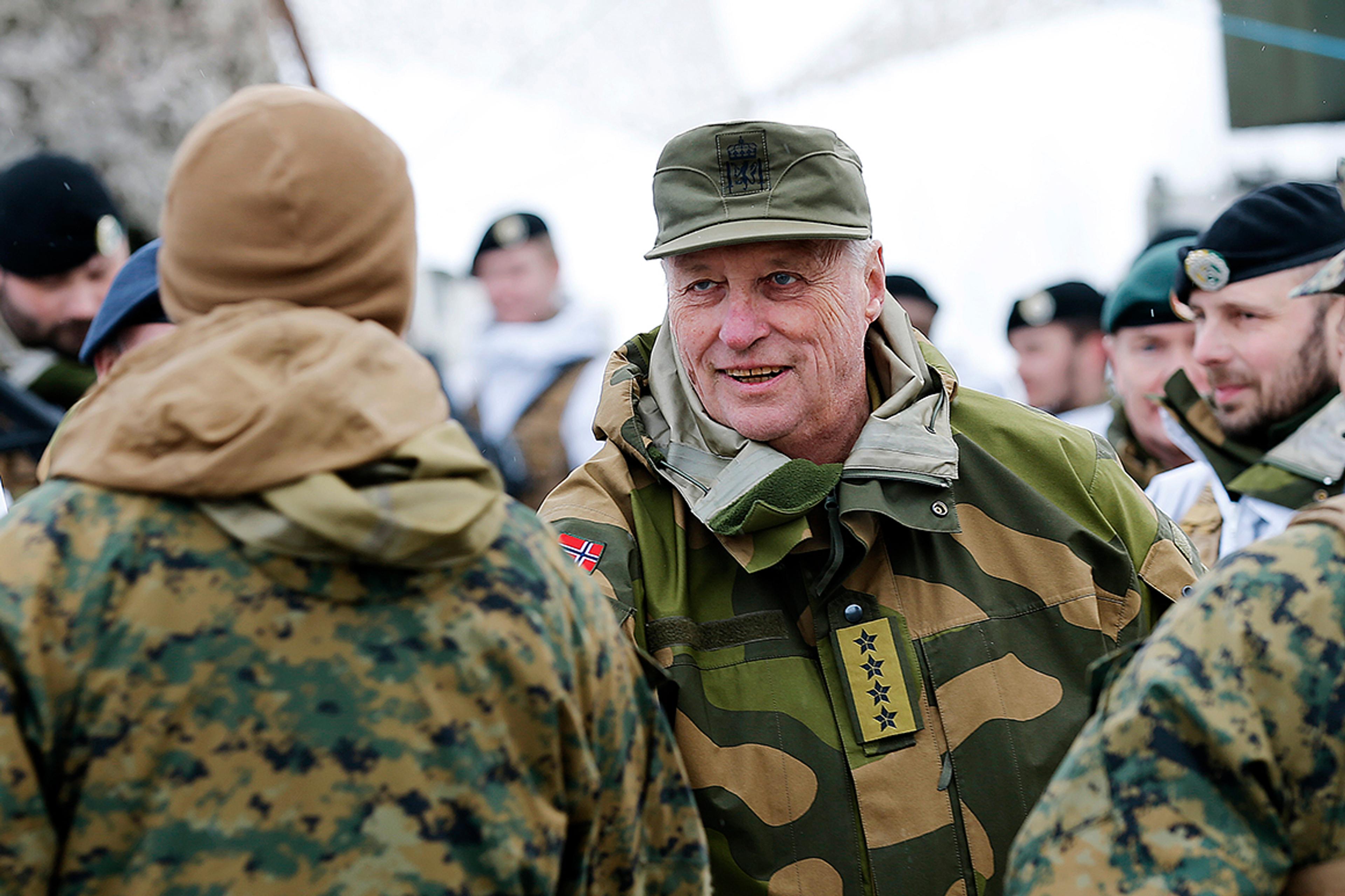 Kongen i feltuniform - firestjernes general - håndhilser på soldat med ryggen til. Kongen smiler. Flere soldater omgir dem.