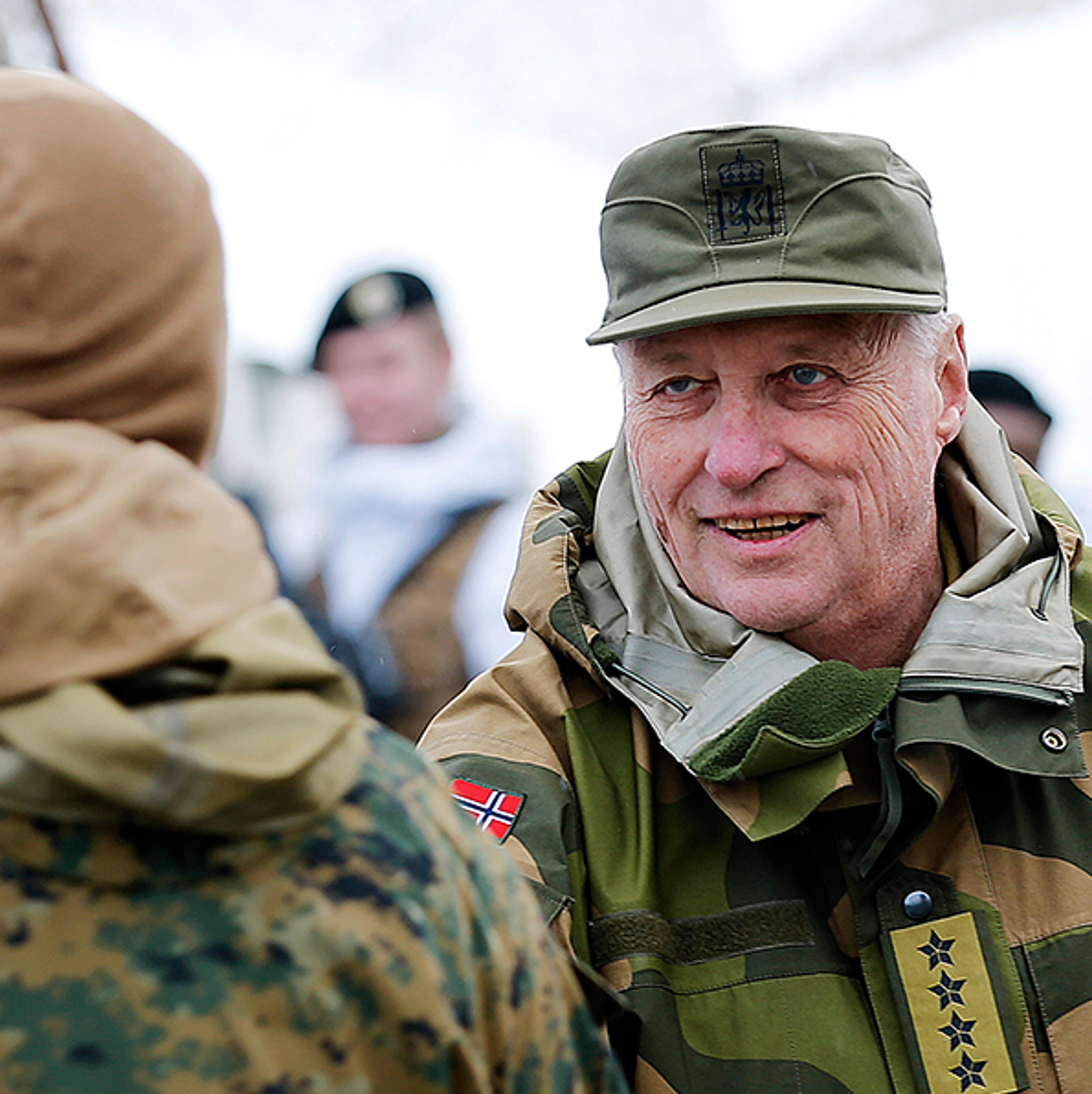 Kongen i feltuniform - firestjernes general - håndhilser på soldat med ryggen til. Kongen smiler. Flere soldater omgir dem.