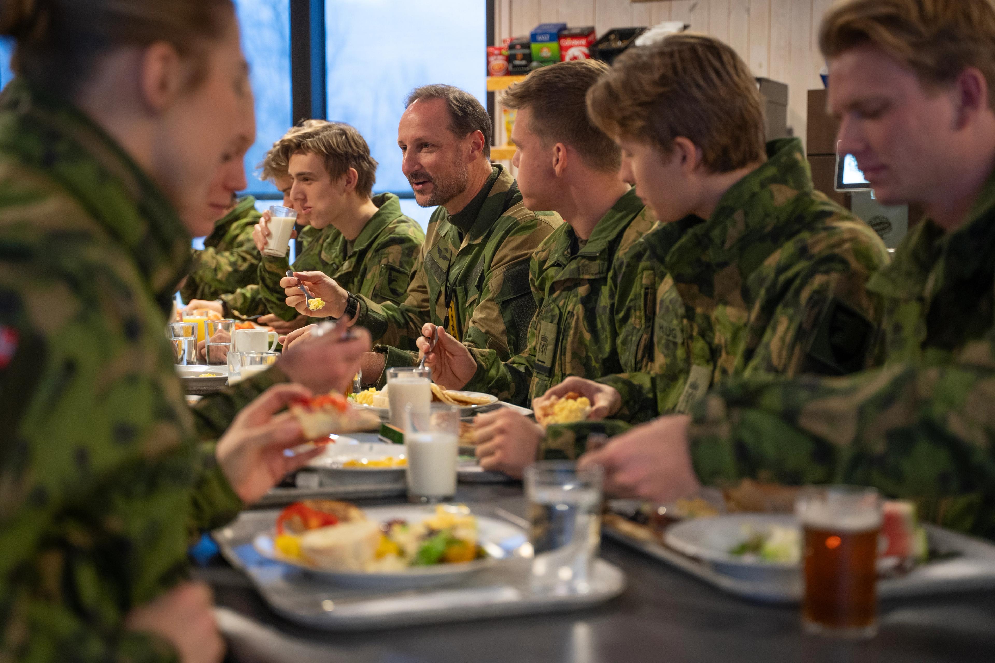 Kongen og soldatene sitter rundt et langbord og spiser frokost fra brett og prater. Alle i uniform.