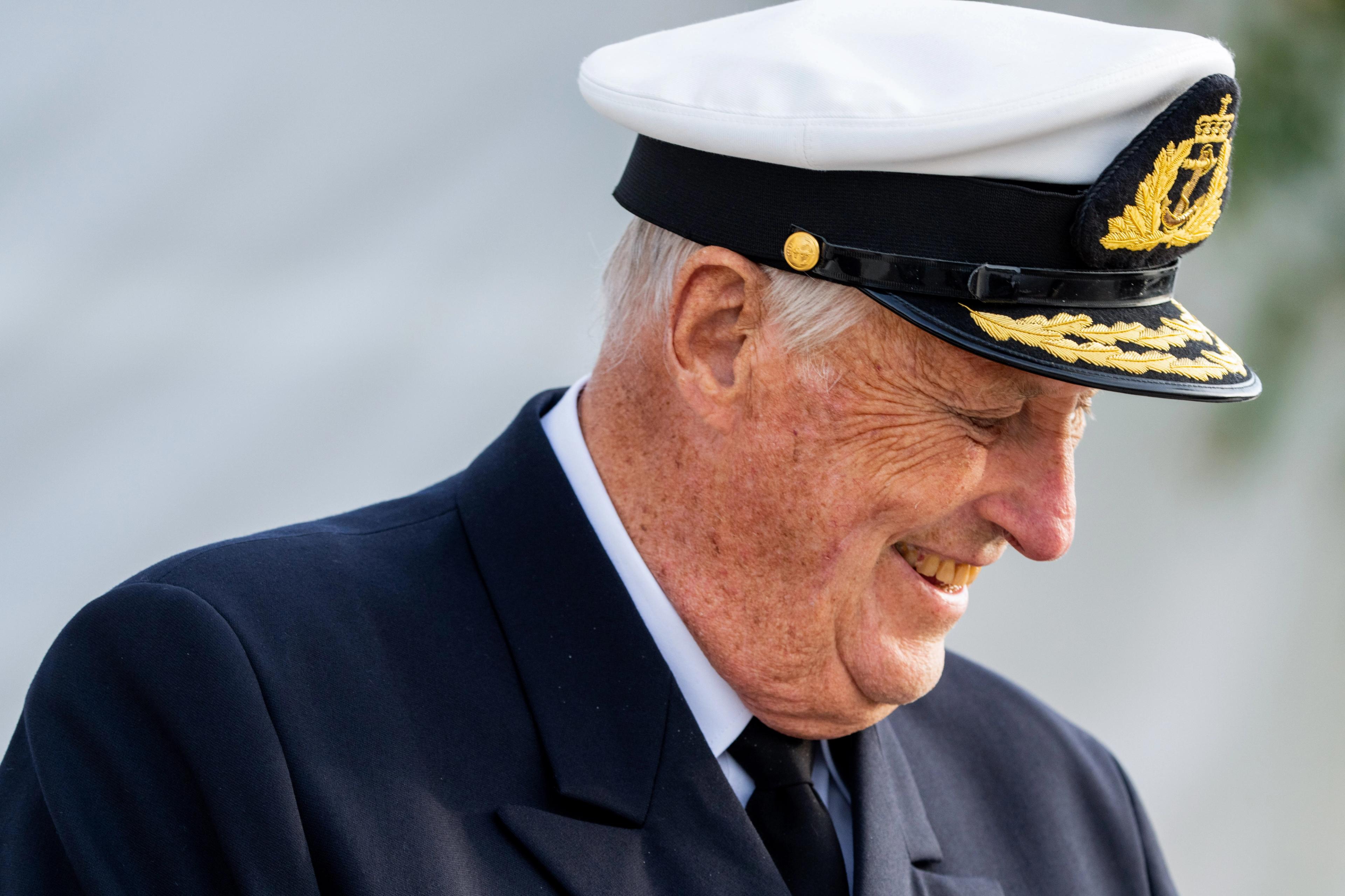 Kong Harald, ikledd Sjøforsvarets uniform, kikker skrått nedover mot bakken mens han smiler.