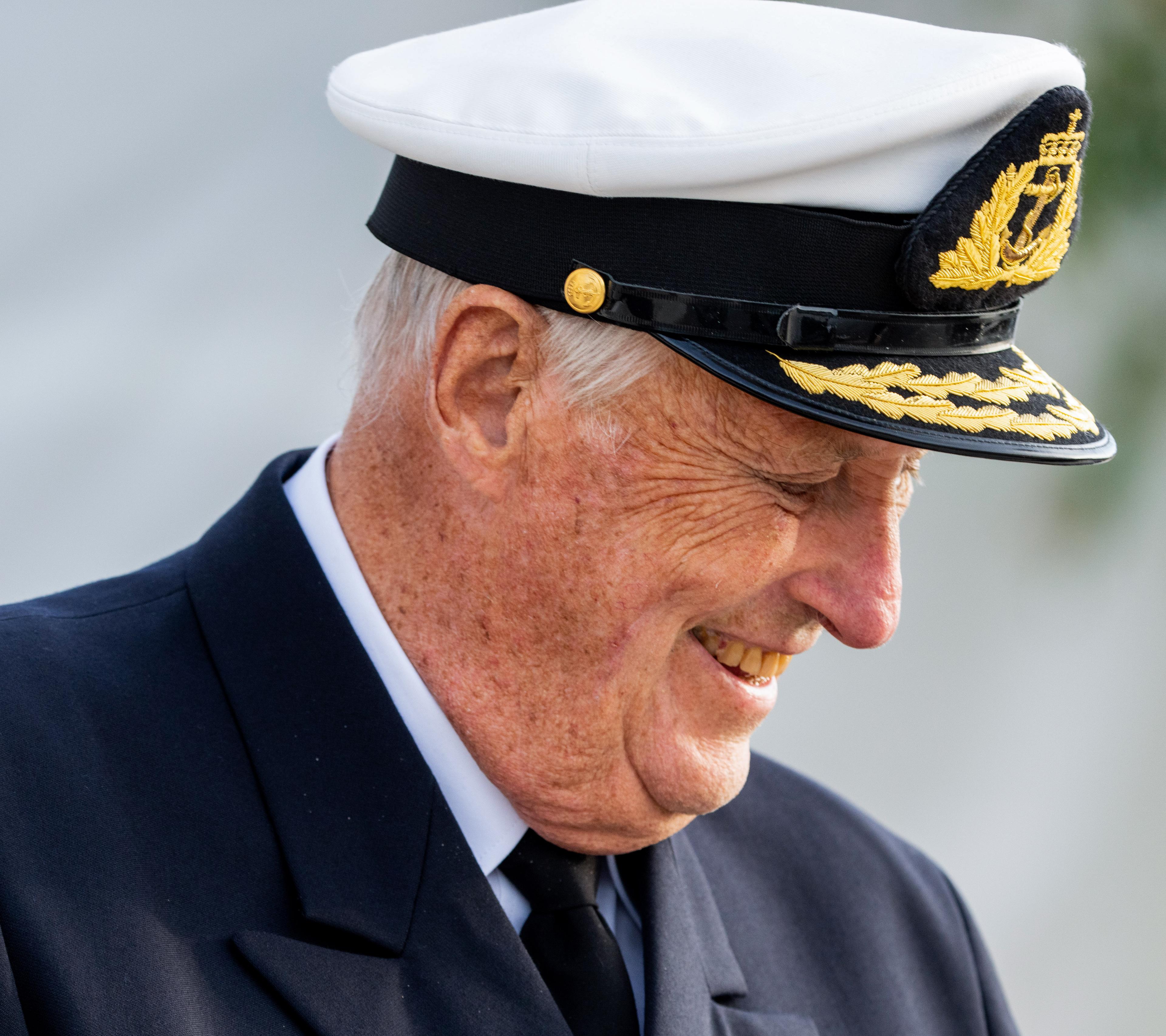 Kong Harald, ikledd Sjøforsvarets uniform, kikker skrått nedover mot bakken mens han smiler.