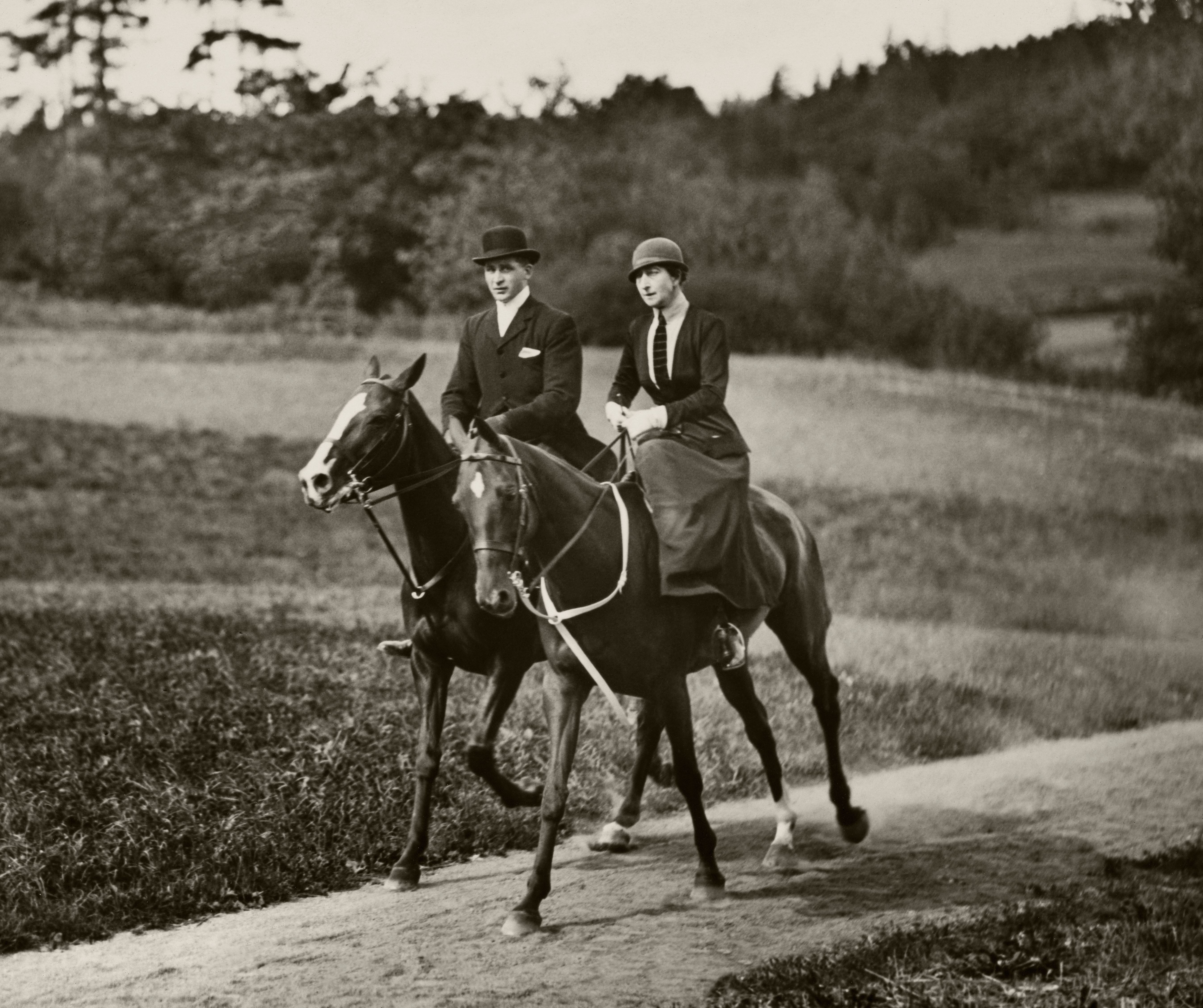 Dronning Maud til hest langs en grusvei i følge med en annen rytter. Begge i rideantrekk, noe som for dronningens del betø sidt skjørt, jakke med slips og hatt. Hun red med sidesal.