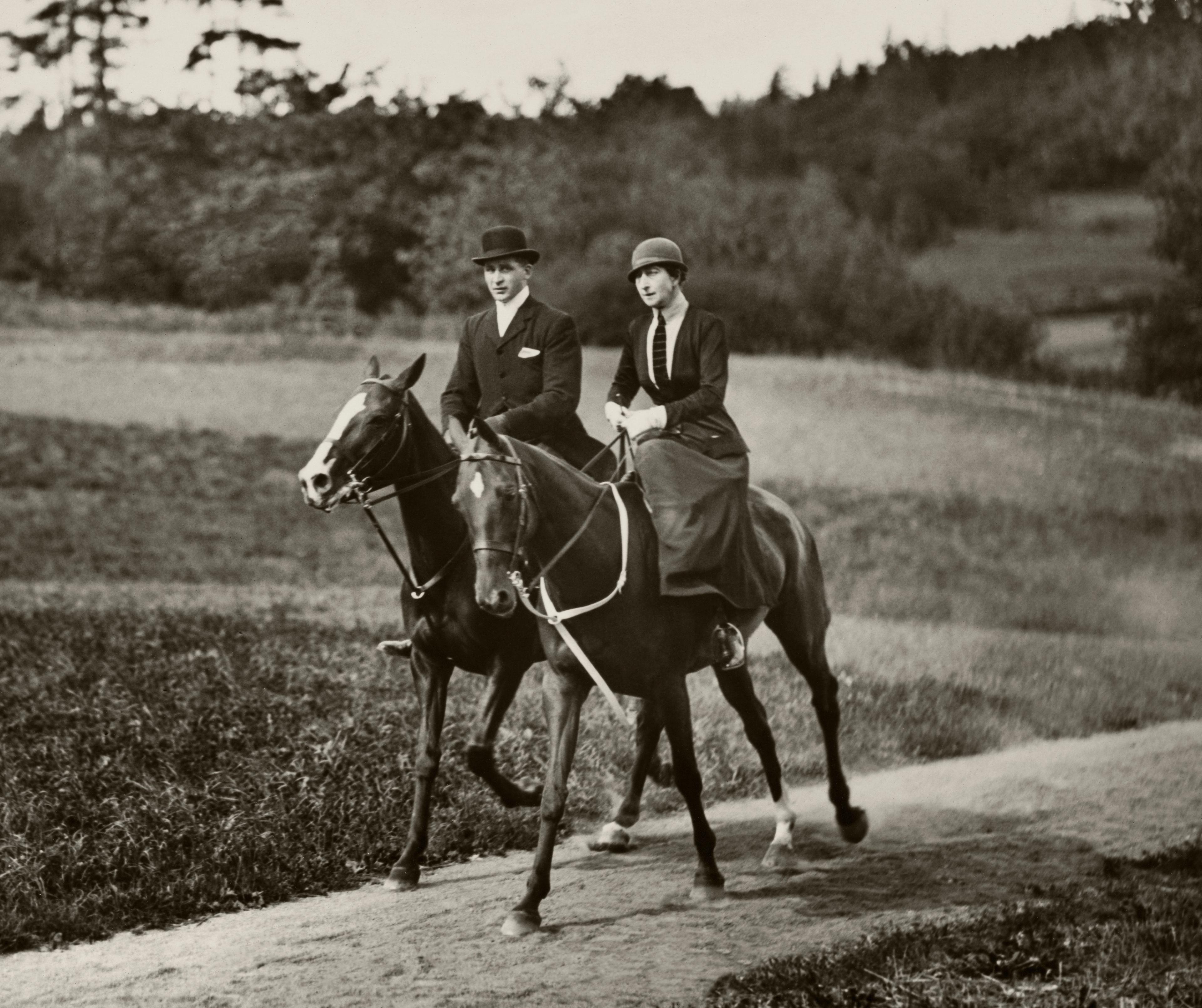 Dronning Maud til hest langs en grusvei i følge med en annen rytter. Begge i rideantrekk, noe som for dronningens del betø sidt skjørt, jakke med slips og hatt. Hun red med sidesal.