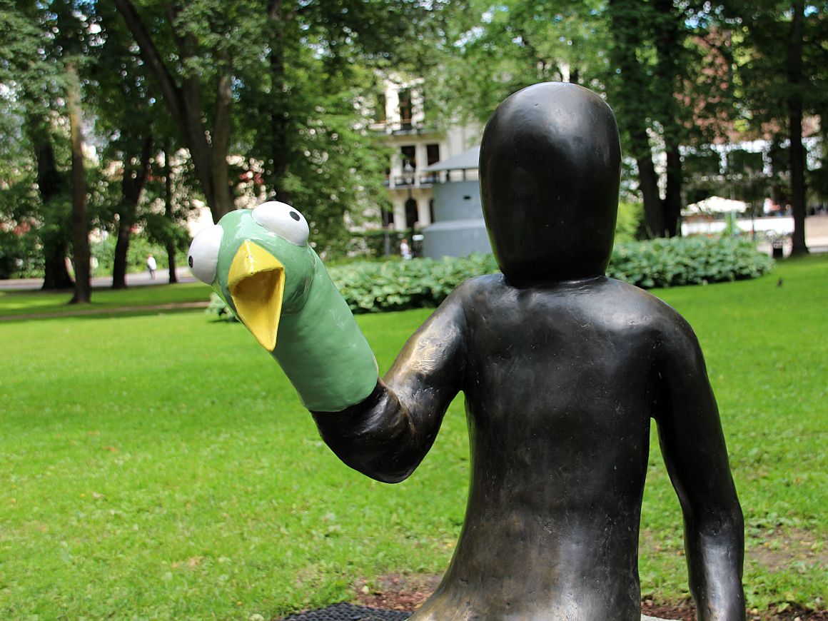 En støpt menneskefigur sitter på en stenblokk. Hodet er uten ansiktstrekk. Et grønt fuglehode med store øyne og åpent, gult nebb er tredd nedover den ene hånden som en dukke.