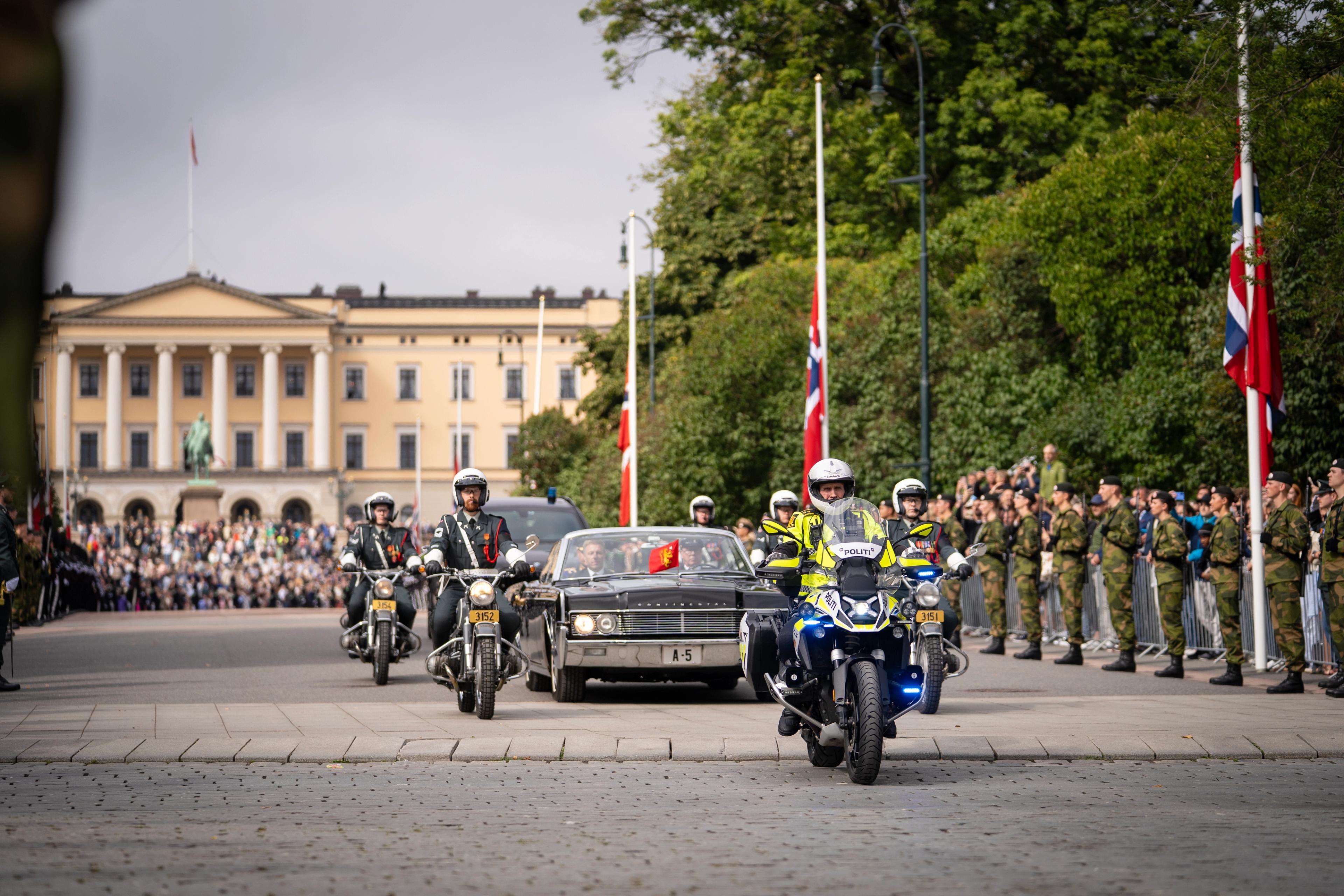 Bilen "A5" i eskorte med flere motorsykler rundt. Kong Haakon sitter i bilen. Slottet sees i bakgrunnen.