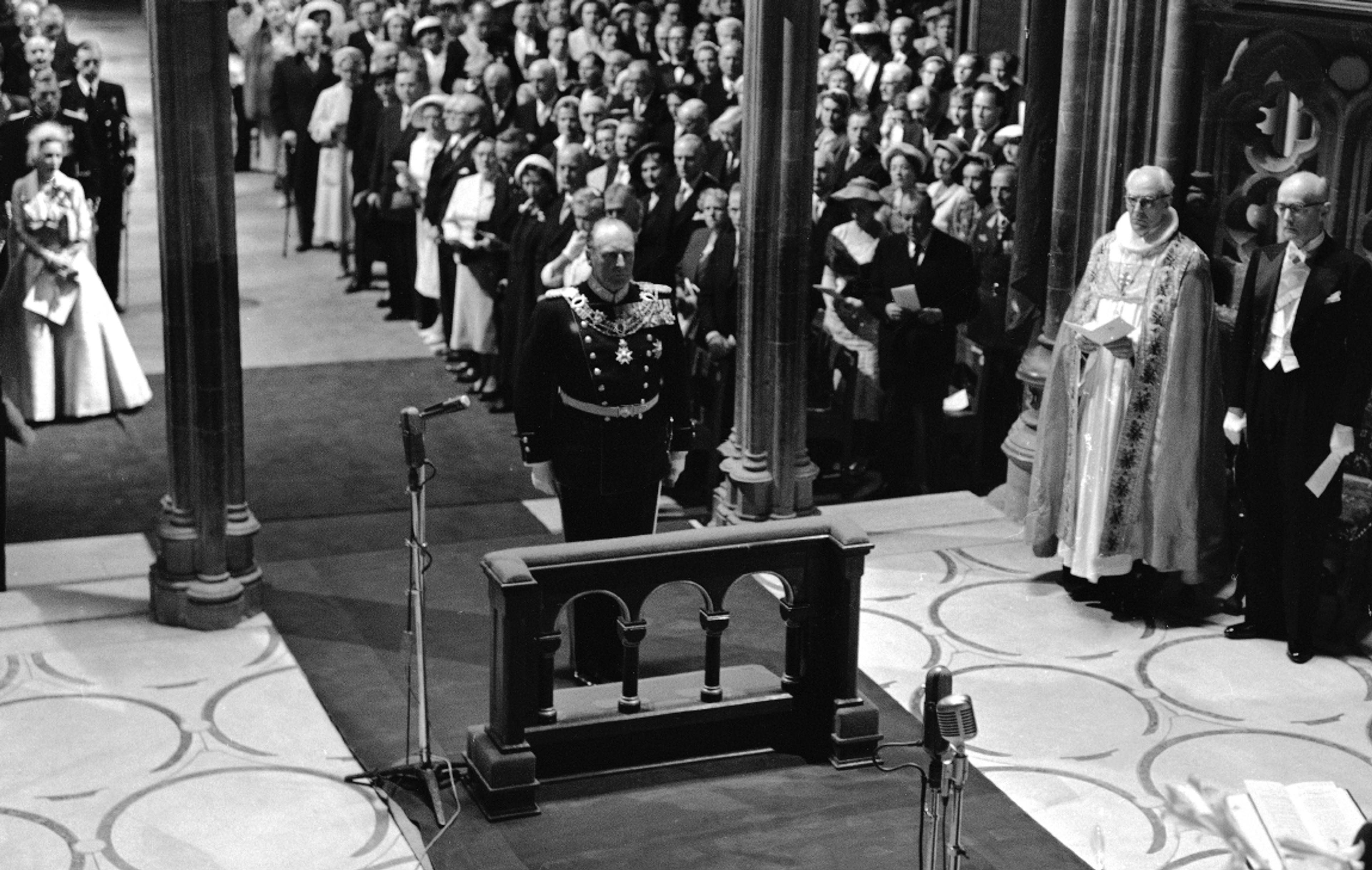 Sort-hvittbilde fra Nidarosdomen. Kong Olav står i gallauniform fremst i kirken og vil snart knele for å motta velsignelsen.