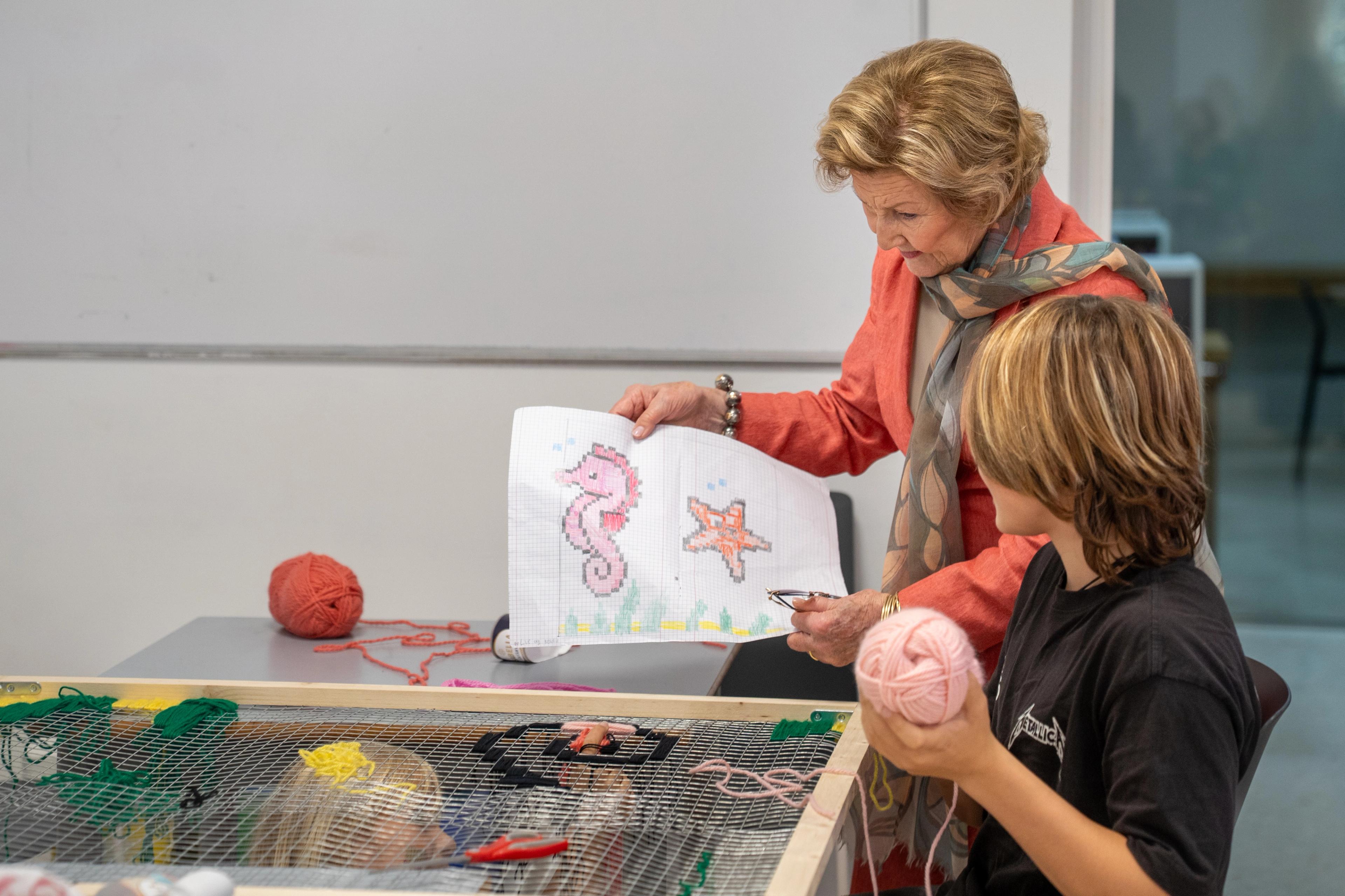 Dronning Sonja held opp eit ark med ein sjøhest og ei sjøstjerne på. Ein ung gut sit framfor ei ramme i treverk, med eit garnnøste i handa.