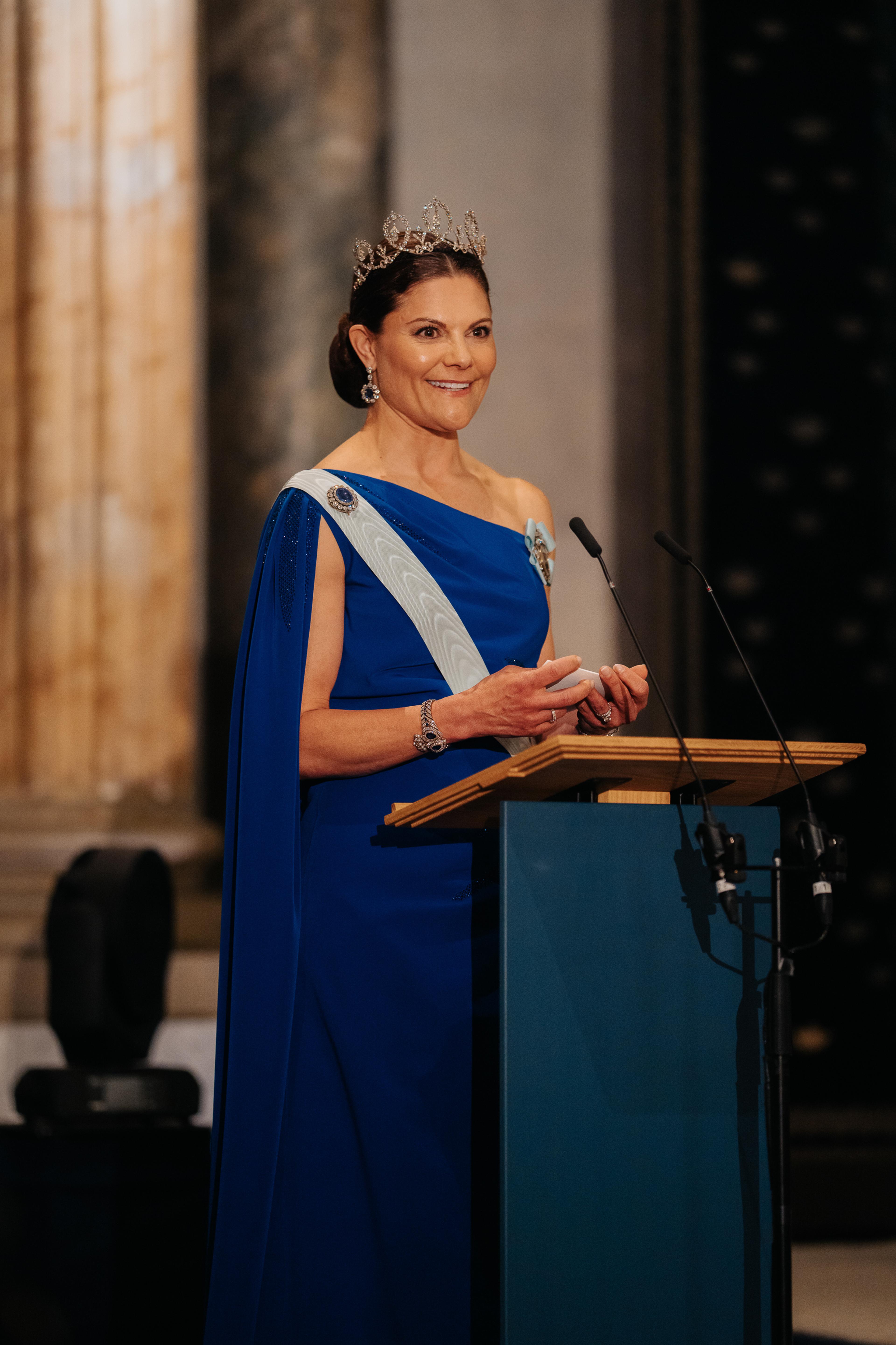 Kronprinsesse Victoria på talerstolen kledd i blå gallakjole, lyseblått ordensbånd og tiara.