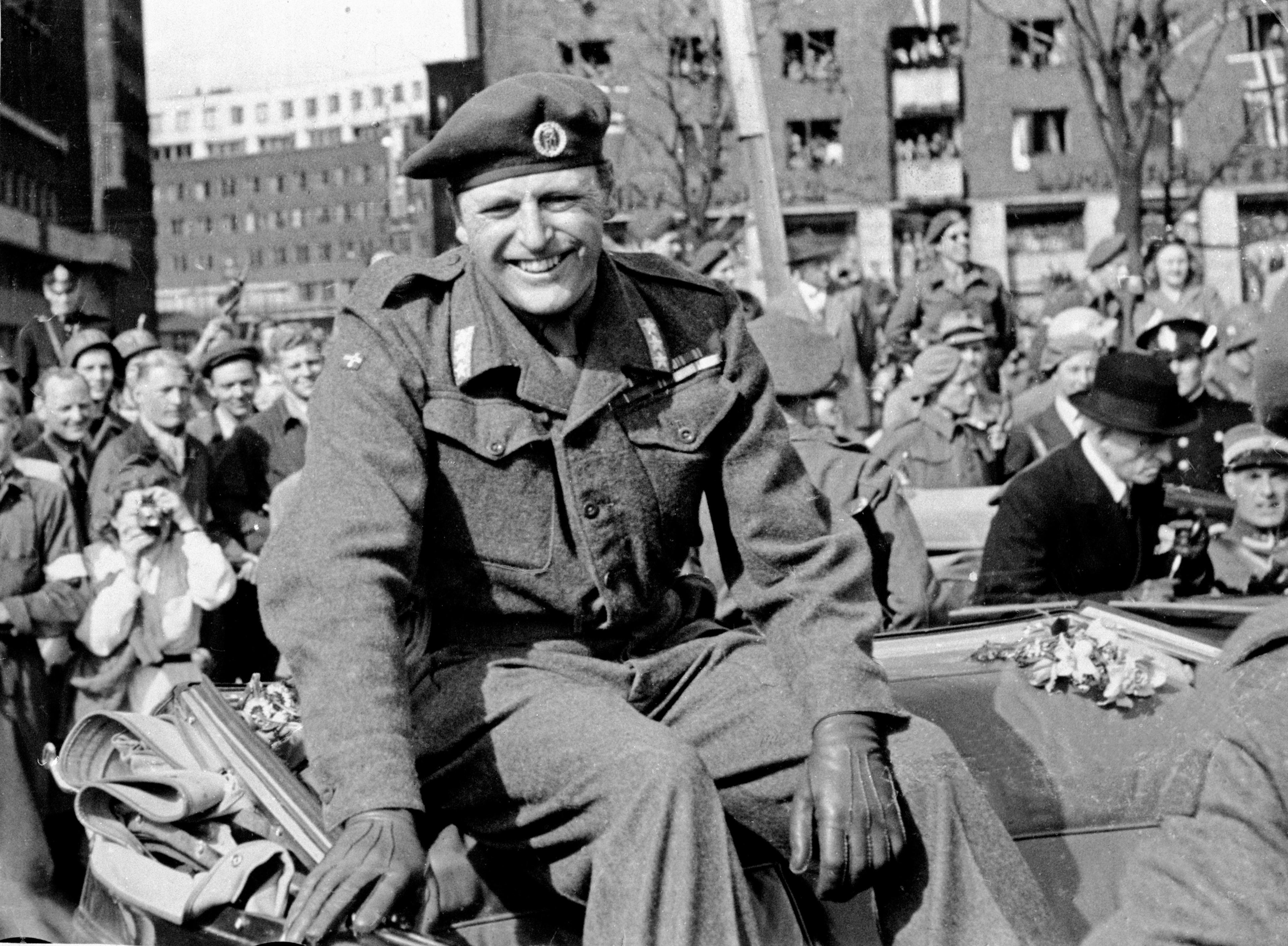 Kronprins Olav i feltuniform sitter med et bredt smil oppå bilens foldede kalesje. Lener seg mot de mange mennskene som omkranser bilen for å hilse.