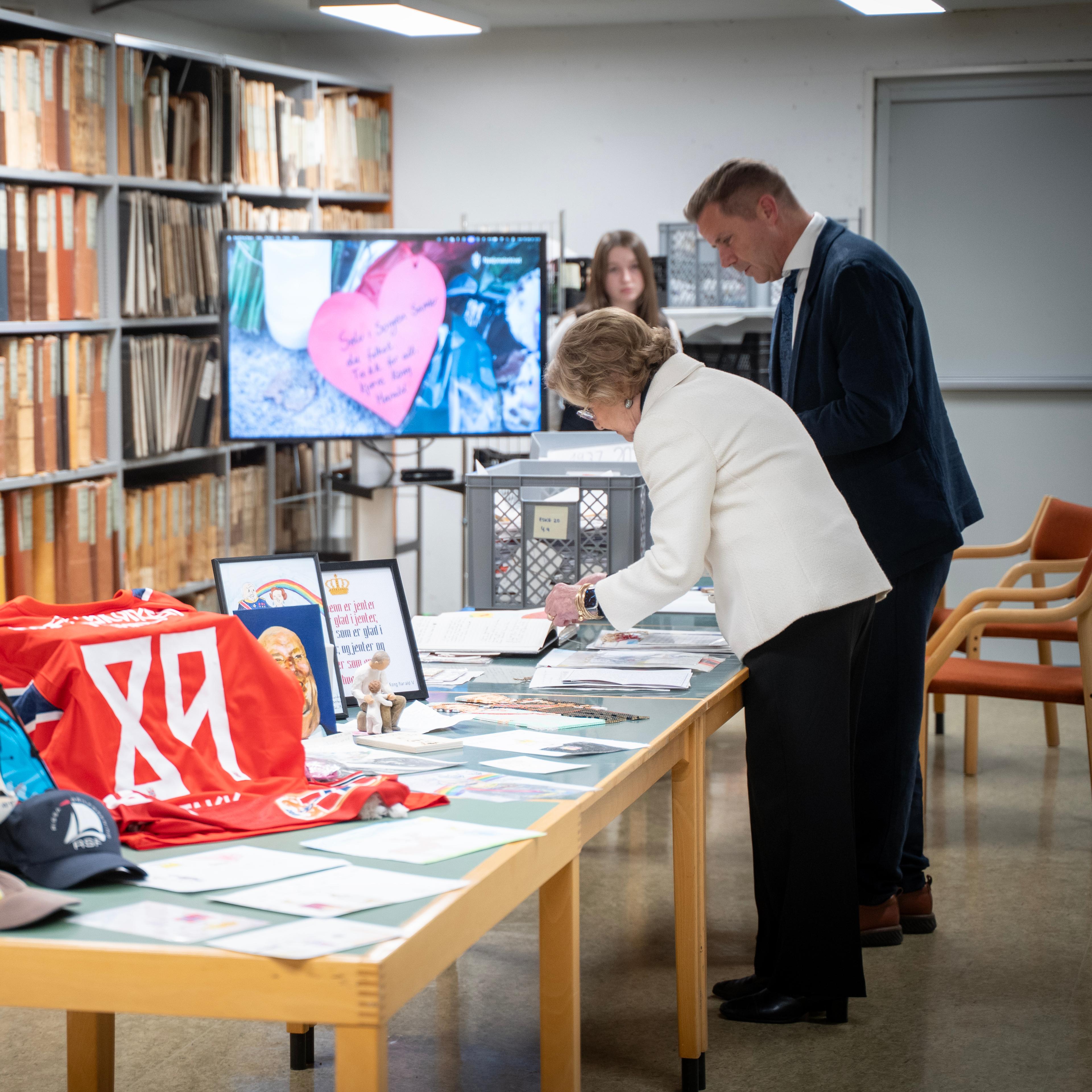 Dronning Sonja og Stian Norli står over et langbord med gjenstander, tegninger, broderier og brev.