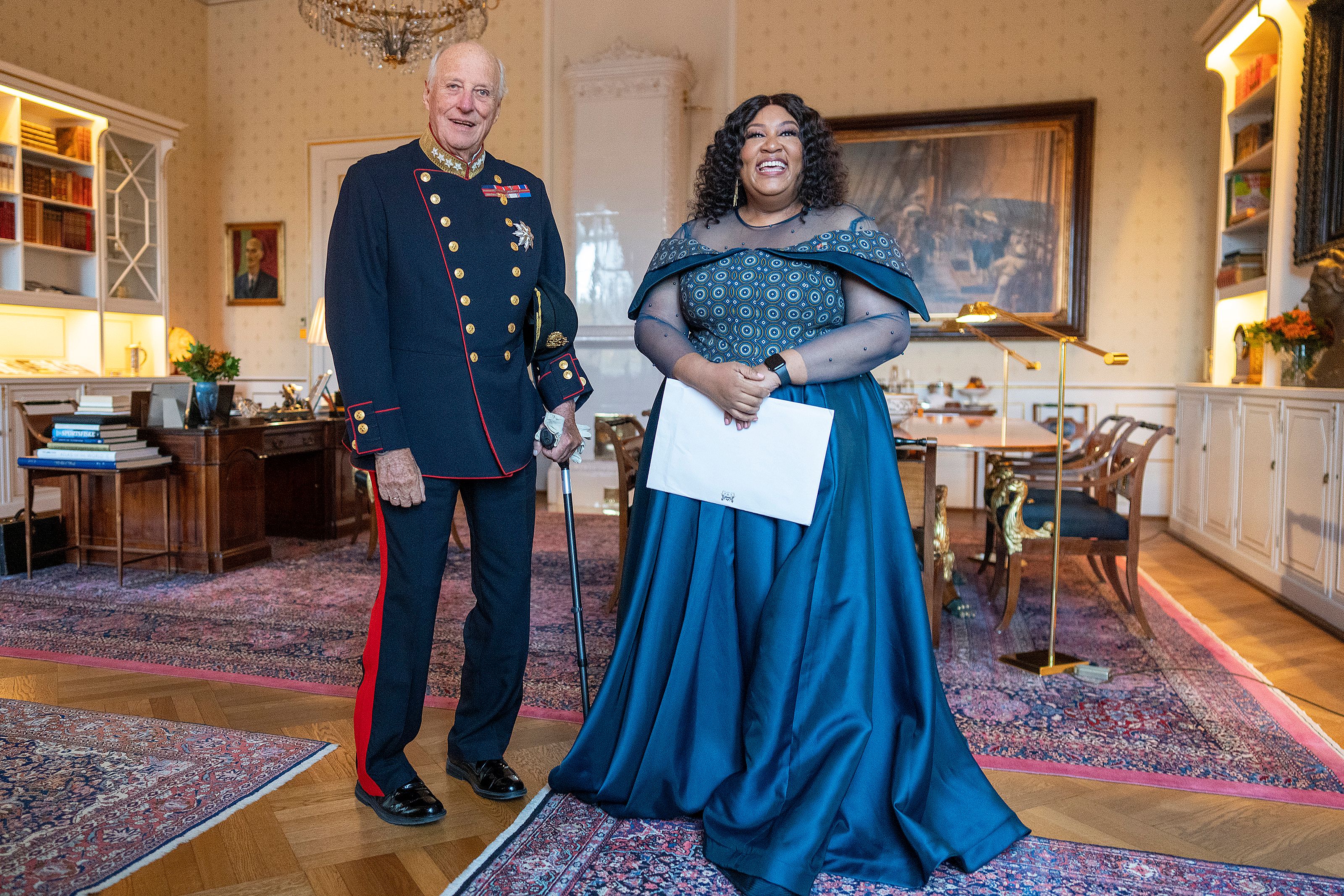 Kongen står smilende i gallauniform ved siden av ambassadøren, som er kledd i fotsid, petroleumsblå kjole. Hun holder en konvolutt med akkreditivene.