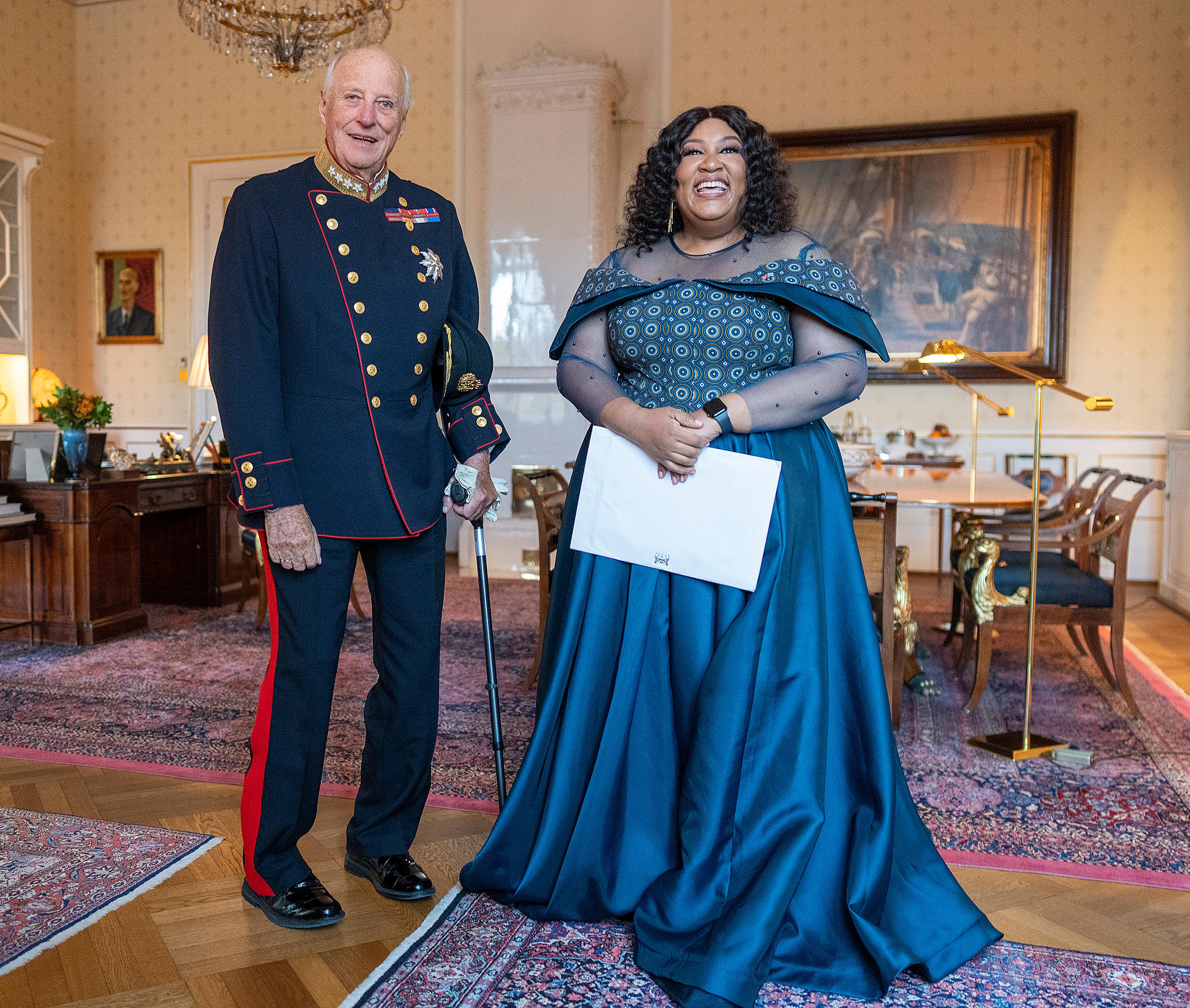 Kongen står smilende i gallauniform ved siden av ambassadøren, som er kledd i fotsid, petroleumsblå kjole. Hun holder en konvolutt med akkreditivene.