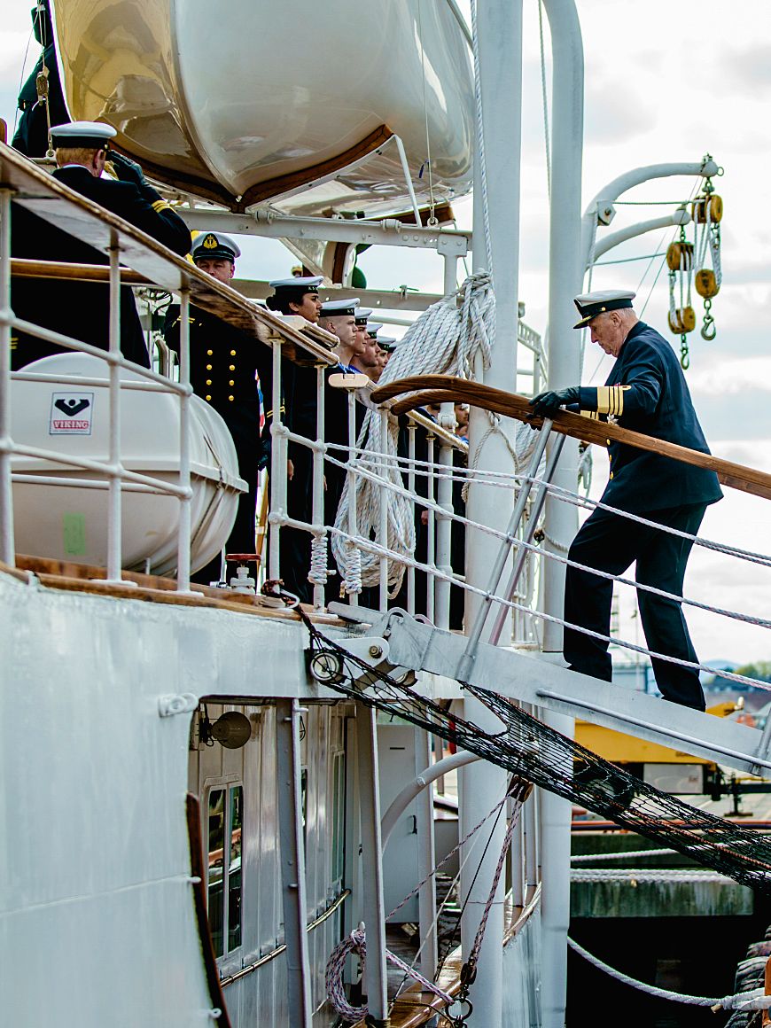 Kongen går opp gangveien fra kaia og om bord i skipet. Han er i marineuniform.