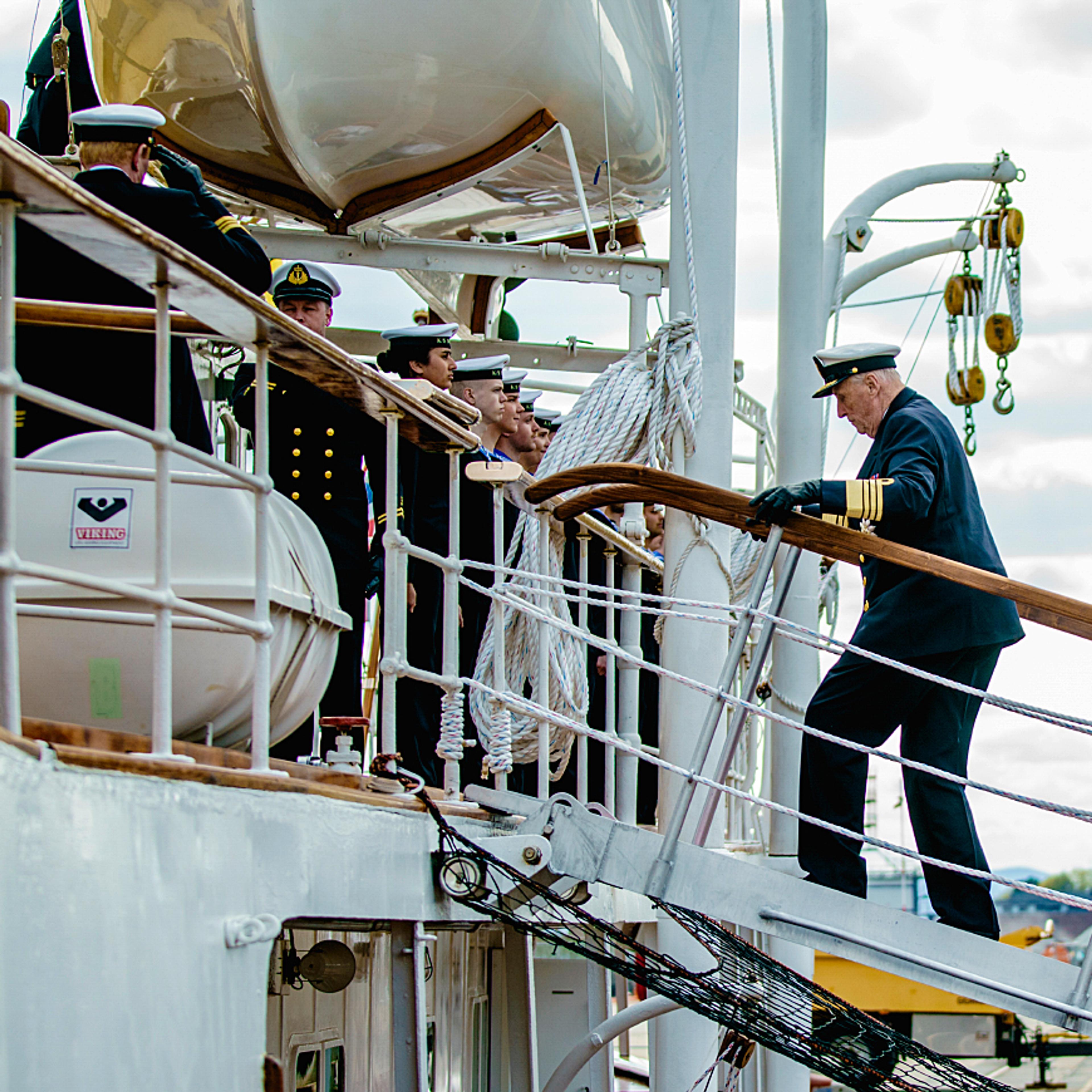 Kongen går opp gangveien fra kaia og om bord i skipet. Han er i marineuniform.