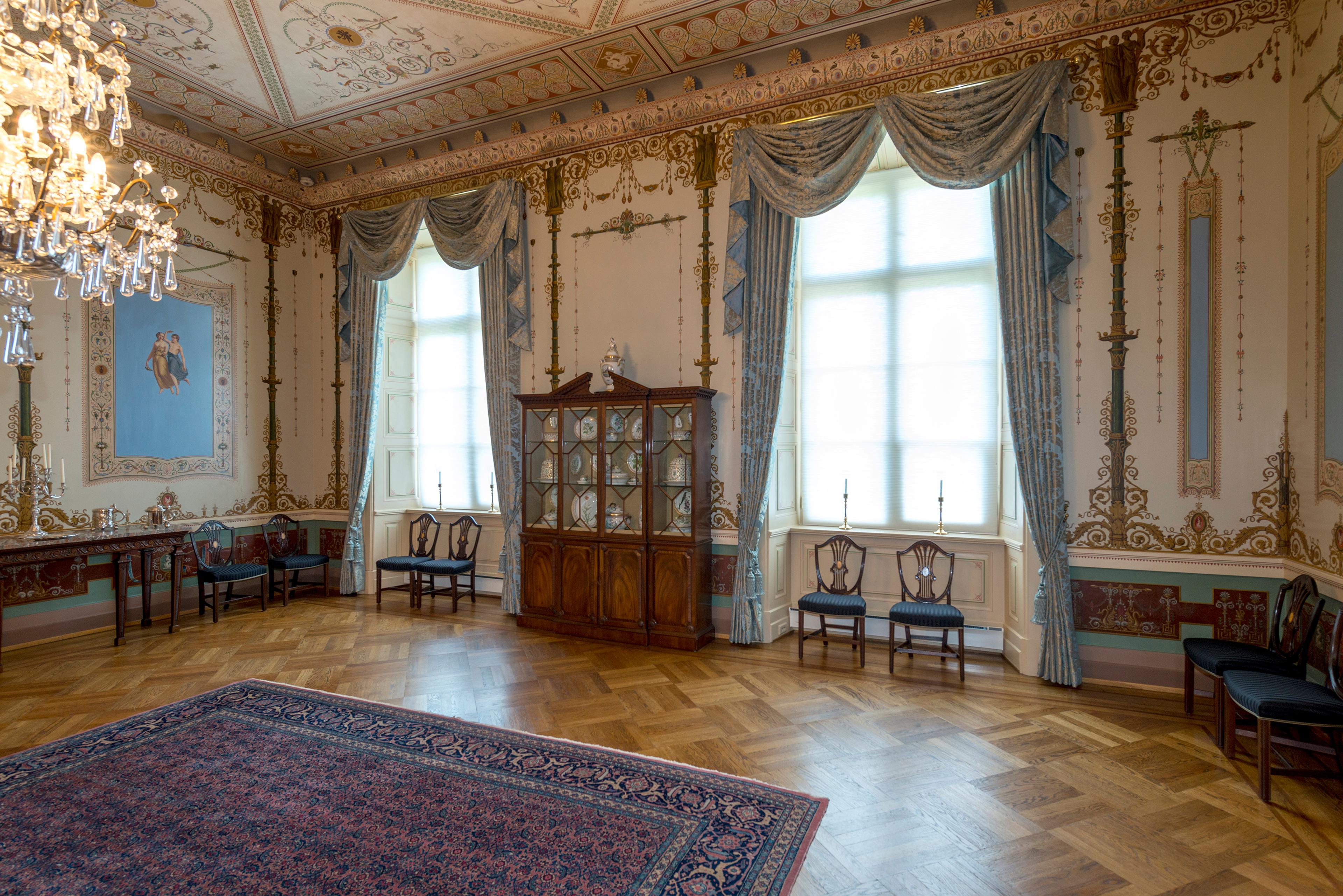 Del av et rom med høye vinduer og rikt dekorerte vegger og tak.