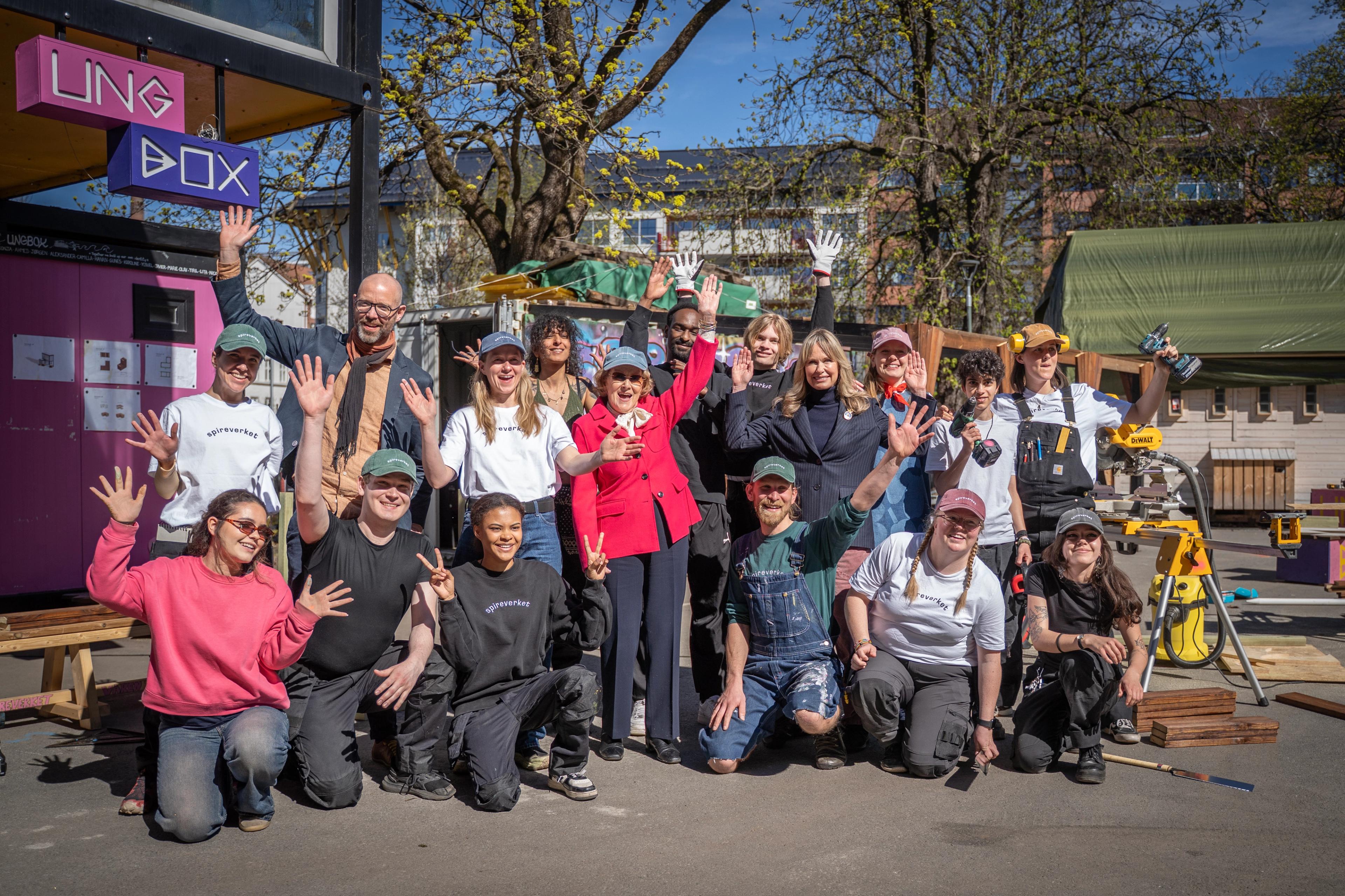 Gruppebilde av Dronning Sonja og ordfører Anne Lindboe sammen med ansatte og unge voksne i Spireverket i arbeidsantrekk. De står ute i strålende solskinn.