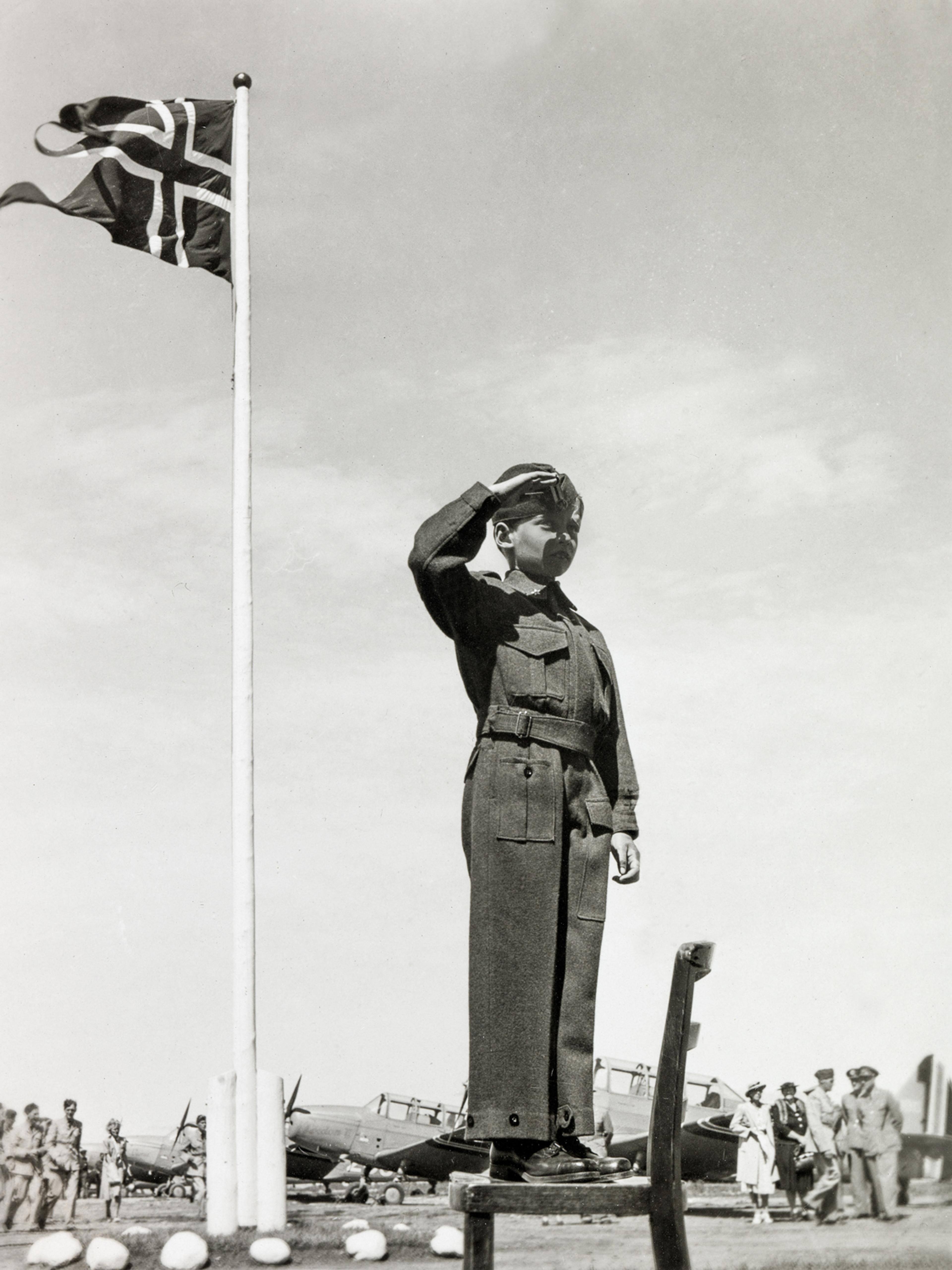 Prins Harald står i en militærlignende uniform og gir honnør til lua. Bak vaier det norske flagget fra en flaggstang.