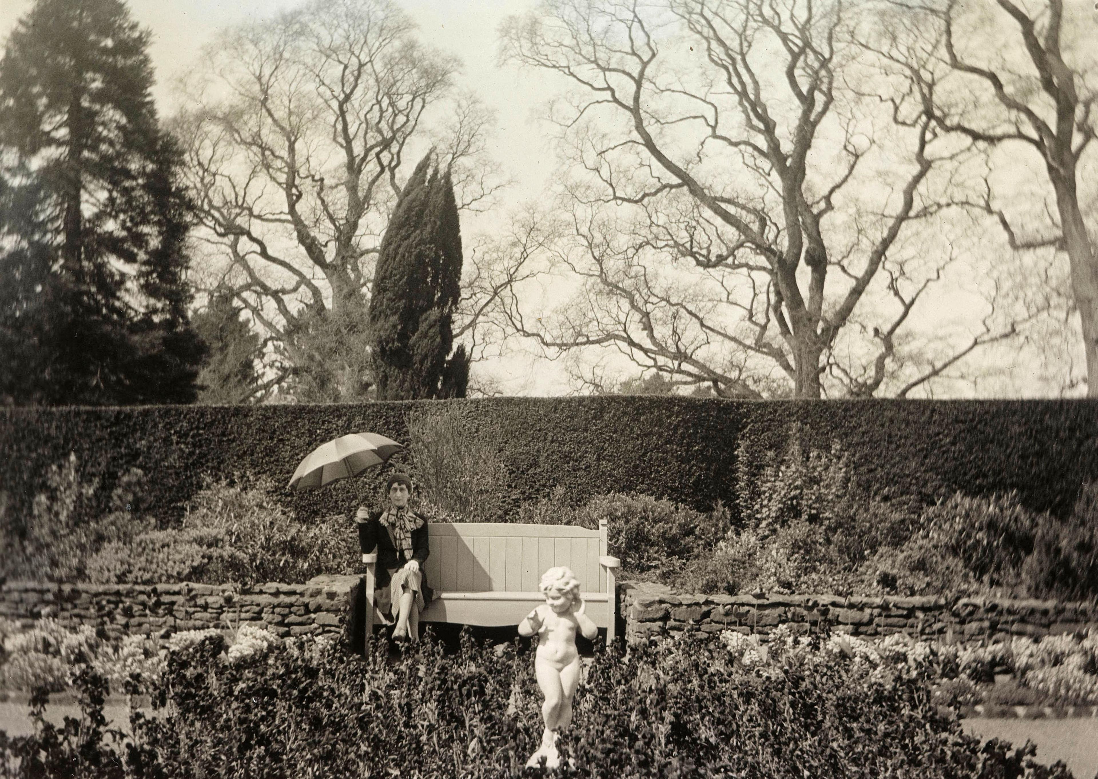 Dronningen sitter med parasoll på en benk og ser mot en liten skulptur i bedet foran.