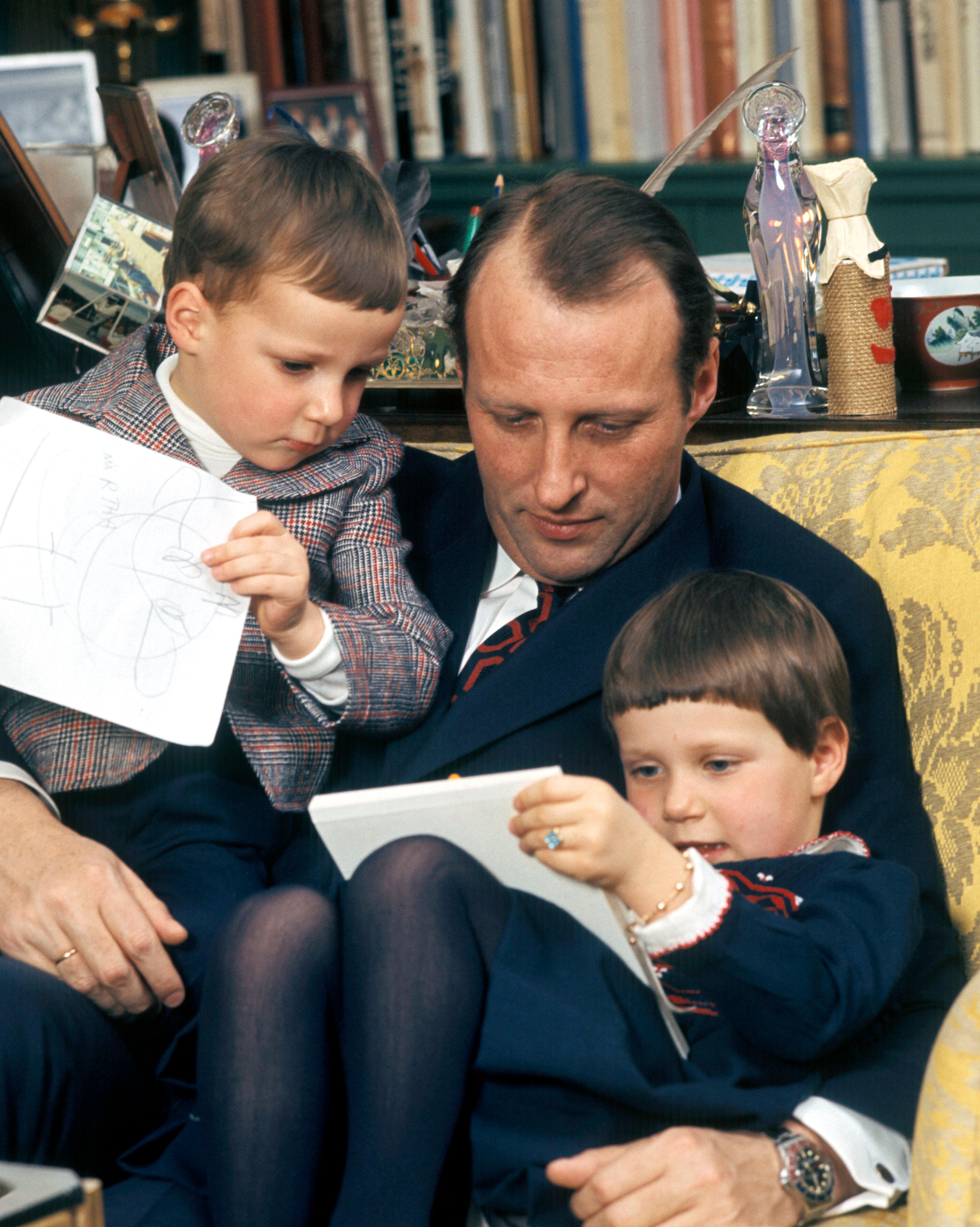 40 år gamle Kronprins Harald sitter i en god stol med de to barna på fanget. Han studerer barnas tegninger.