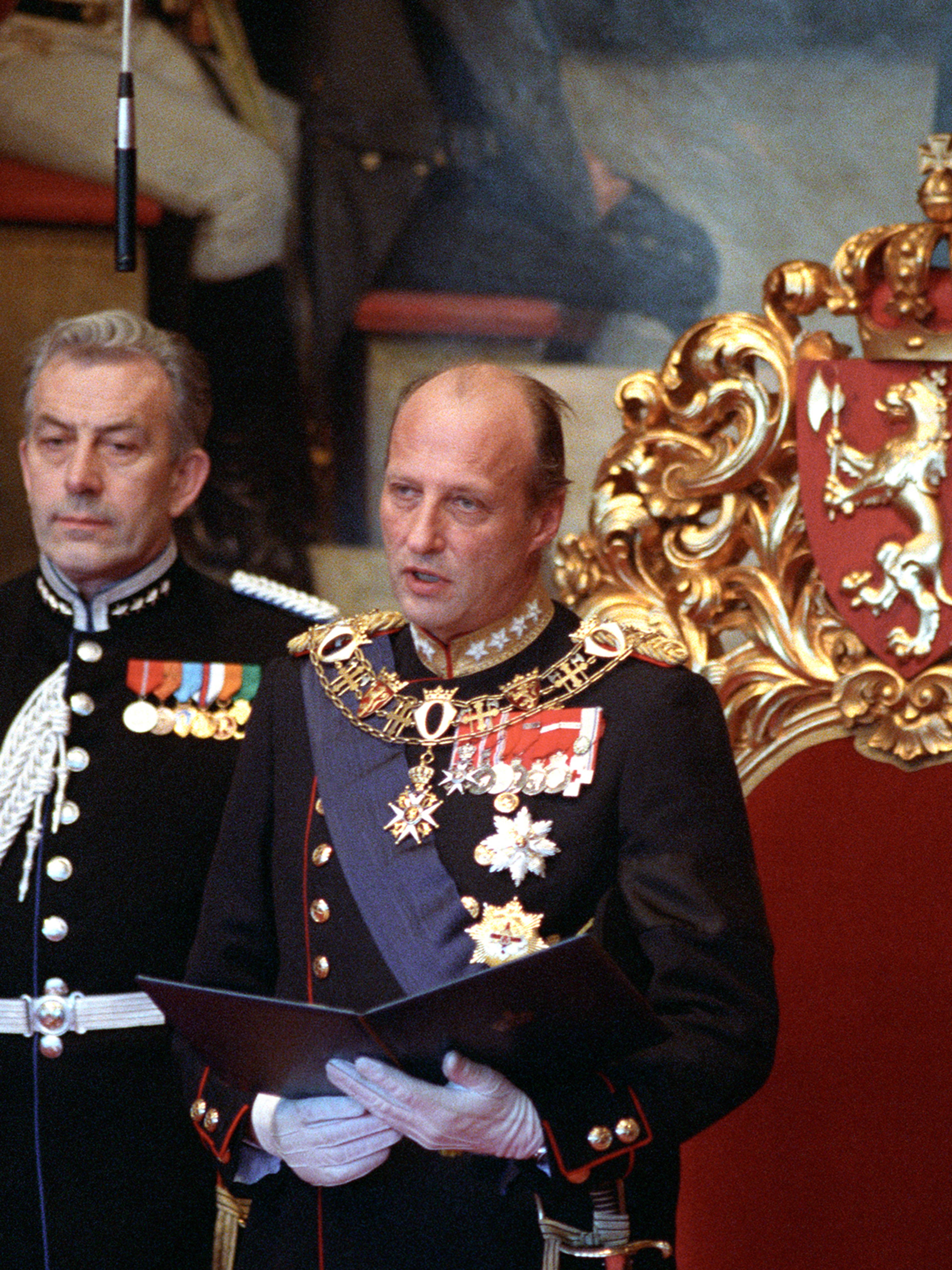 Kong Harald står foran tronstolen i rødt og gull, og han leser fra en oppslått skinnmappe. Han er ikledd Hærens gallauniform.