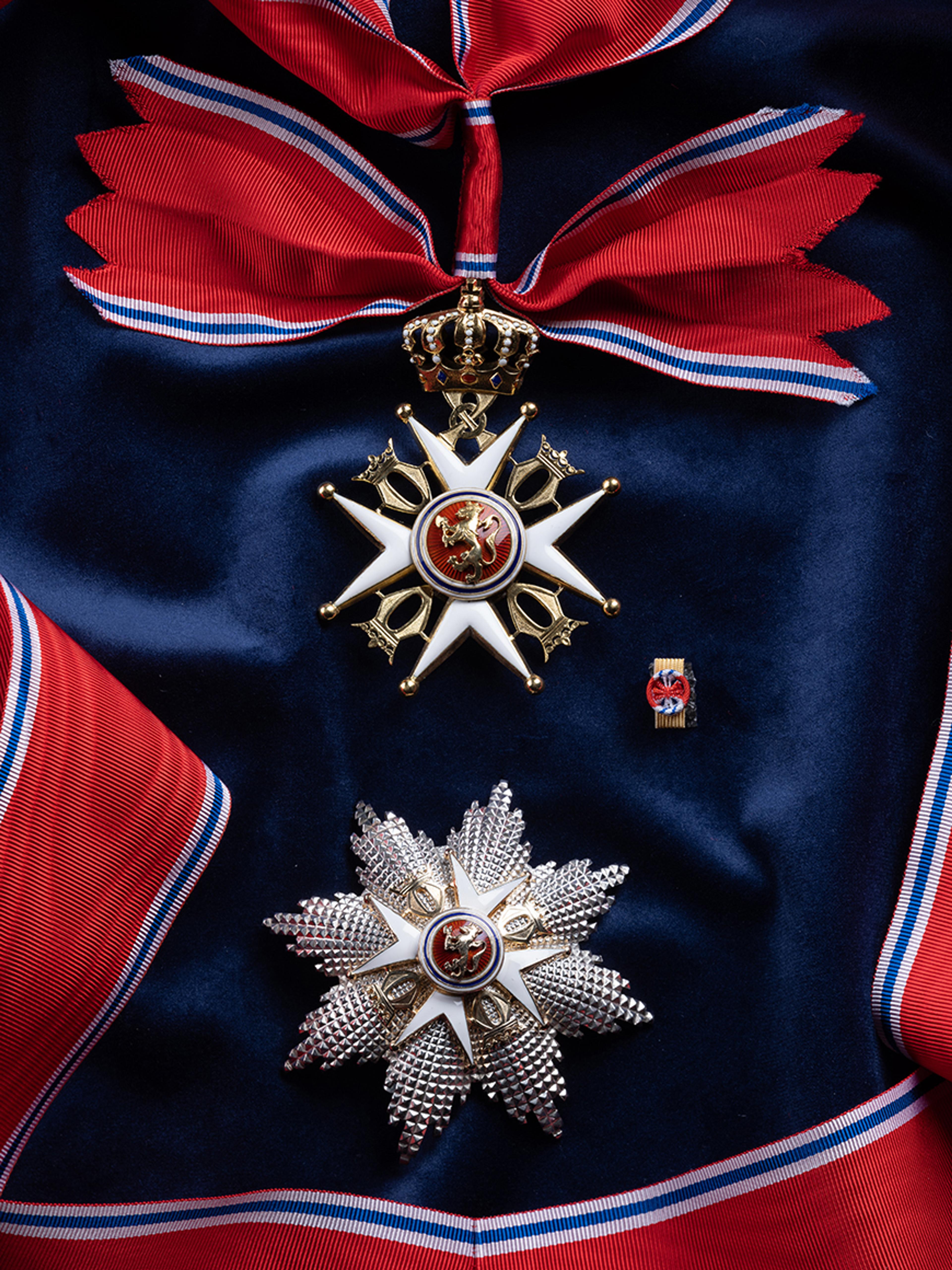 Nærbilde av ordensmerket festet i rødt bånd med blå og hvite striper på en bakgrunn av blått klede. Ordenens krasjan og button ligger ved siden av.
