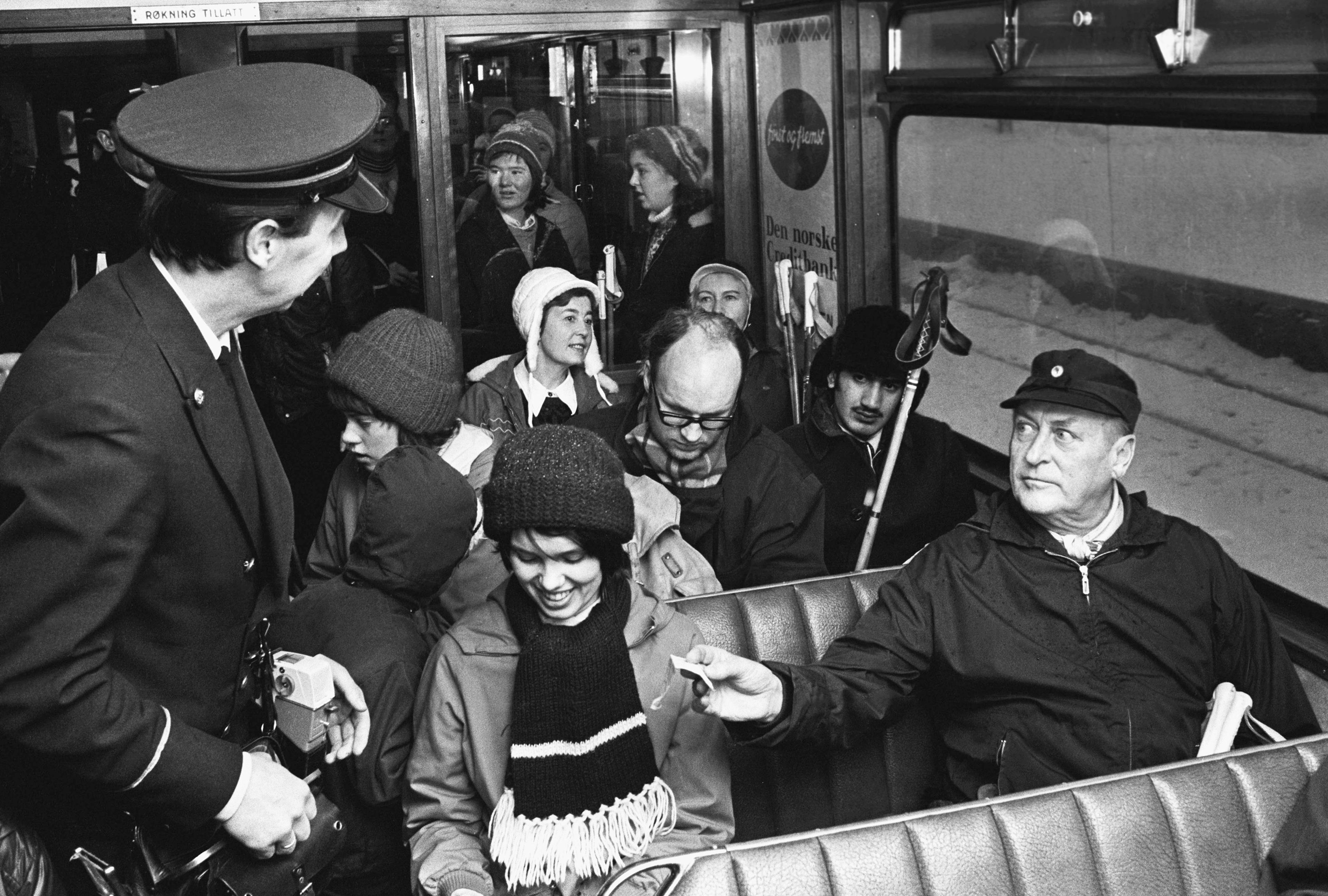 Kong Olav sitter på trikken med alle andre. Rekker fram peker til konduktøren for å betale for billetten.