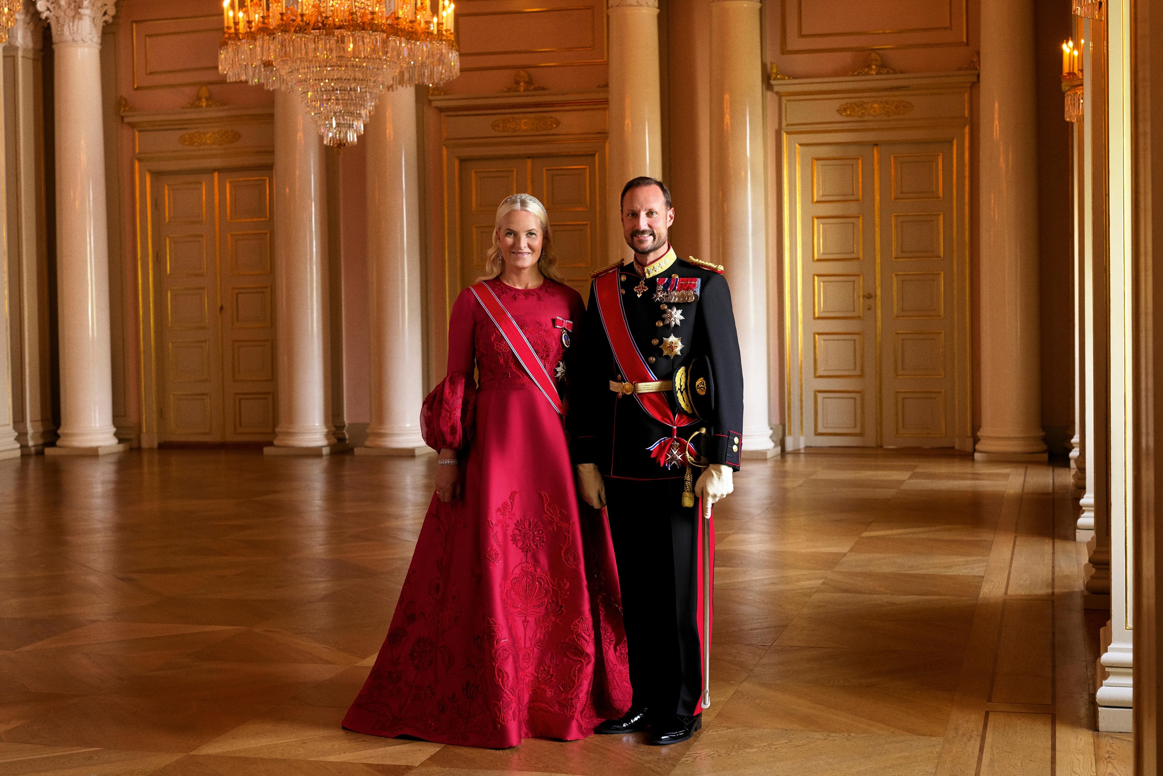 Kronprinsparet i helfigur i Store festsal. Kronprinsen har gallauniform med ordener og medaljler, Kronprinsessen har rød gallakjole og ordensbånd.